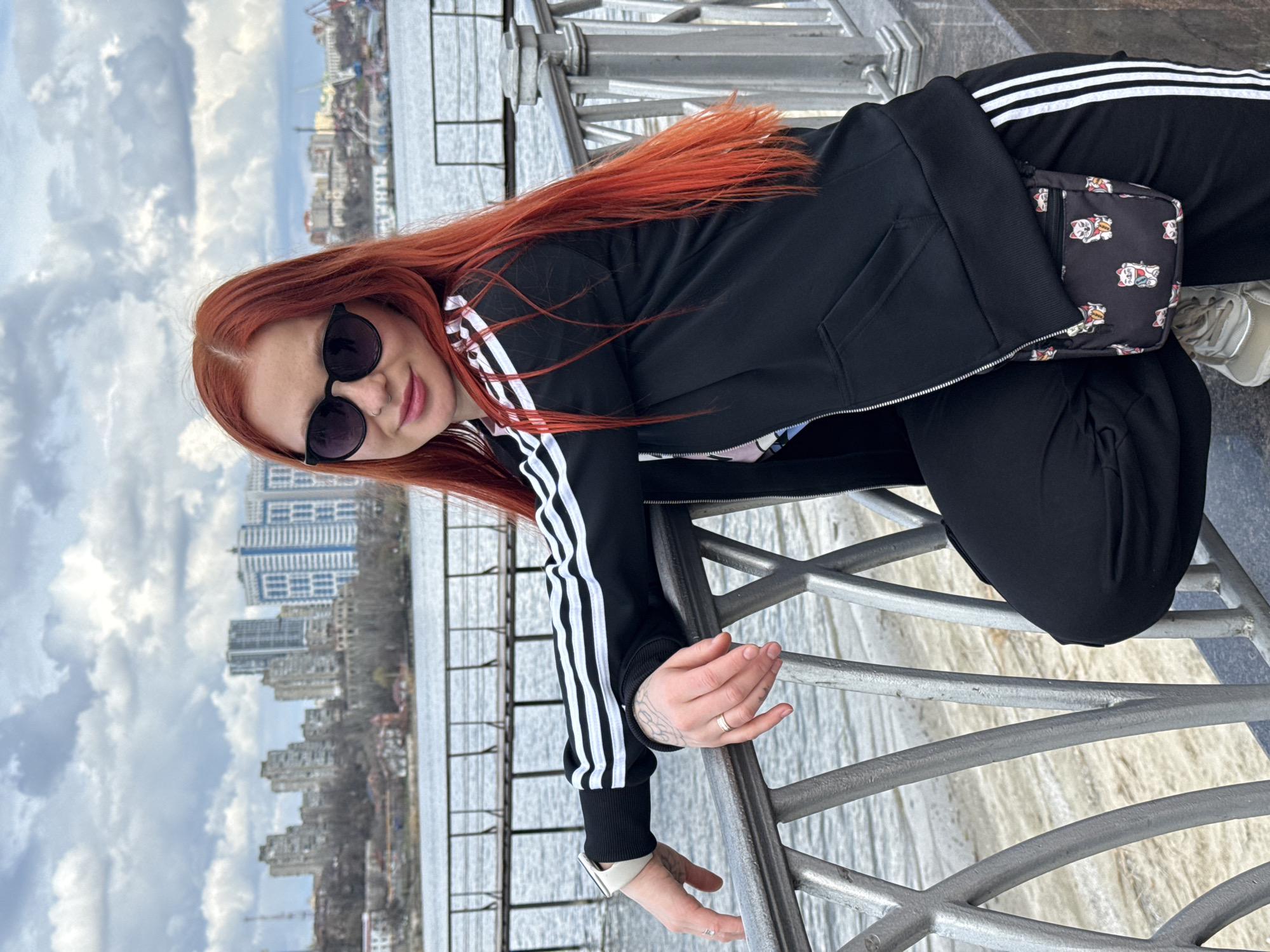 A young woman with long, red hair stands near a metal railing in Rostov-on-Don, Russia. She wears sunglasses, a black Adidas jacket with white stripes, and black pants. A small, patterned purse hangs at her hip. In the background, the Don River and city buildings are visible under a cloudy sky. The woman is leaning towards the railing, one hand resting on it.