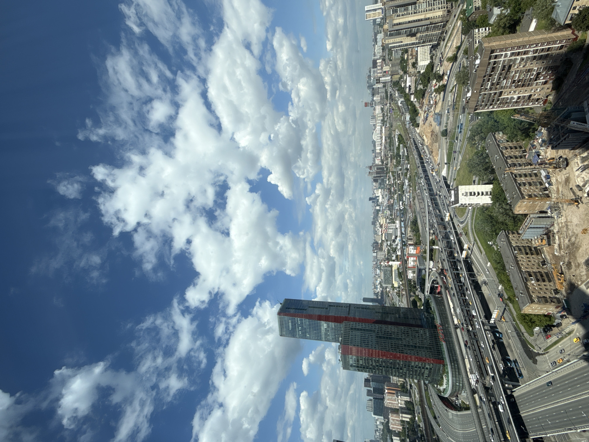 This media file presents a high-angle view of a sprawling urban landscape, seen from an elevated perspective. The image is oriented in portrait mode, with a vast expanse of blue sky and scattered white cumulus clouds dominating the left side. The right side and central portion of the frame showcase a dense city environment, characterized by a complex network of multi-lane highways and elevated roadways, visible with numerous vehicles indicating moderate traffic flow.
A prominent, modern skyscraper with a distinctive facade featuring dark panels and striking red horizontal stripes stands tall in the lower-left-mid section of the urban area, appearing to reach into the cloud-filled sky. Surrounding this landmark and the extensive road system are a variety of other buildings, ranging from high-rise residential blocks to commercial structures and older apartment buildings, some of which are under active construction, evidenced by cranes, scaffolding, and excavated ground. Green trees and patches of vegetation are interspersed throughout the concrete jungle.
The scene is brightly lit by what appears to be midday or early afternoon sun, suggesting clear weather conditions with excellent visibility. Faint text, including "КОНТАКТ" (KONTAKT) and "ШИНЫ" (SHINY - Tires), can be discerned on a white roadside sign. The image captures the dynamic and expansive nature of a modern metropolitan area, with the location identified as Khimki, Russia.