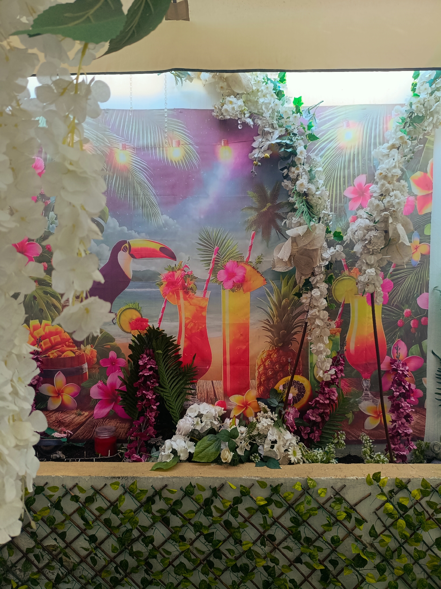 The image displays a tropical-themed backdrop at a wedding reception in Rafelbunyol, Spain. The backdrop features a beach scene with palm trees, a toucan, tropical drinks, pineapples, and mangoes. Artificial white, pink, and purple flowers, along with green foliage, are arranged in front of the backdrop, creating a lush and festive atmosphere. The foreground shows a wall covered with a lattice of branches and artificial green leaves. The overall scene suggests a celebration with a vibrant, summery ambiance.