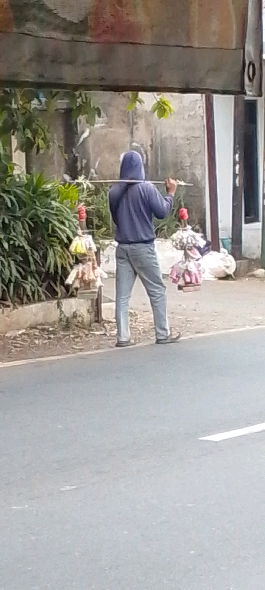 A person wearing a blue hooded jacket and light-colored pants is walking along a paved road. They are carrying a long wooden pole horizontally across their shoulders, with an object or objects hanging from each end of the pole. To the left of the person, there is a dense green bush and some sort of decorative stand with colorful hanging elements. Behind the person, there is a concrete wall and a building. The road appears to be in a semi-urban or rural setting, with vegetation and a somewhat weathered overhead structure visible in the background. The image captures the individual in motion, walking away from the camera. The location is identified as Cilaku, Indonesia.