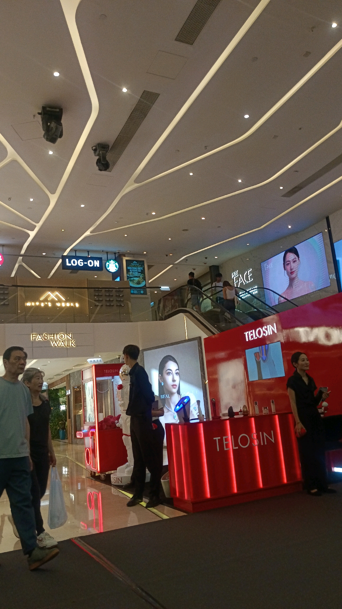 The image captures a scene within a modern indoor shopping mall, likely in Hong Kong, China, given the presence of a Starbucks sign and what appears to be a "Fashion Walk" branding, a known shopping district in Causeway Bay, Hong Kong. In the foreground, several people are visible. On the left, a man and a woman are walking. Behind them, another woman is carrying a white bag. In the center, two men in dark clothing stand near a red counter. To the right, a woman in a black outfit stands near the counter.
The environment is characterized by a high, modern ceiling with intricate white lighting structures. Several large digital screens are displayed. One screen shows the "TELOSIN" logo and advertisements for beauty products, featuring a model. Another screen, further back and higher up, displays the "LOG-ON" store sign and a Starbucks logo. Escalators are visible in the background, with people using them to move between levels. The overall ambiance suggests a busy, upscale retail environment.