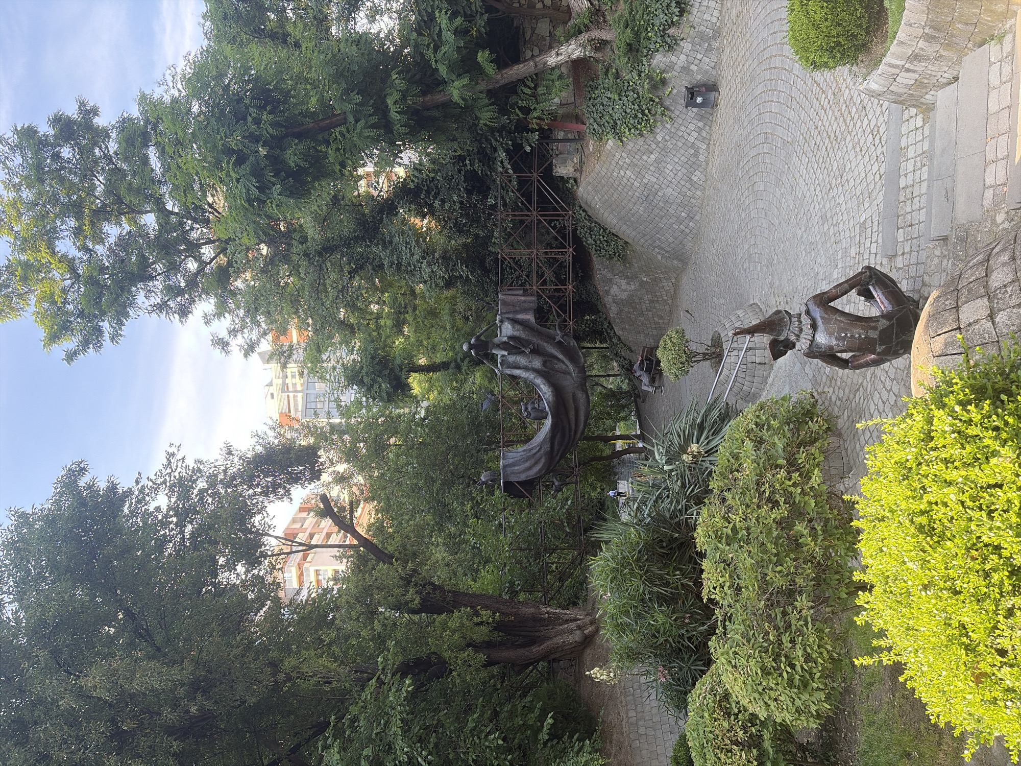 This is a medium shot of a courtyard in Tbilisi, Georgia, bathed in the bright light of what appears to be late afternoon or early evening. The scene is dominated by lush greenery, with mature trees and manicured bushes framing a paved area. The paving stones are laid in a decorative, circular pattern, suggesting a well-maintained public space or garden.
In the foreground, a bronze sculpture depicts a human-like figure with its head bowed, appearing to be in a state of contemplation or distress. The sculpture is situated on a stone base, and a small, neatly trimmed bush grows beside it. Further back, another, more abstract sculpture, possibly made of dark metal, is partially obscured by scaffolding. This sculpture has a flowing, draped form, with smaller figures visible within it.
Beyond the sculptures and greenery, the background reveals buildings, one of which has a modern, multi-story facade with balconies. The presence of these buildings, combined with the style of the paving and the overall atmosphere, suggests an urban courtyard setting, possibly within a historical or cultural district of Tbilisi. The sky is a clear, pale blue with faint wisps of clouds, indicating fair weather. There are no visible text elements in the image. The scene evokes a sense of tranquility and artistic appreciation, with the sculptures adding a layer of thoughtful human presence to the natural beauty of the courtyard.