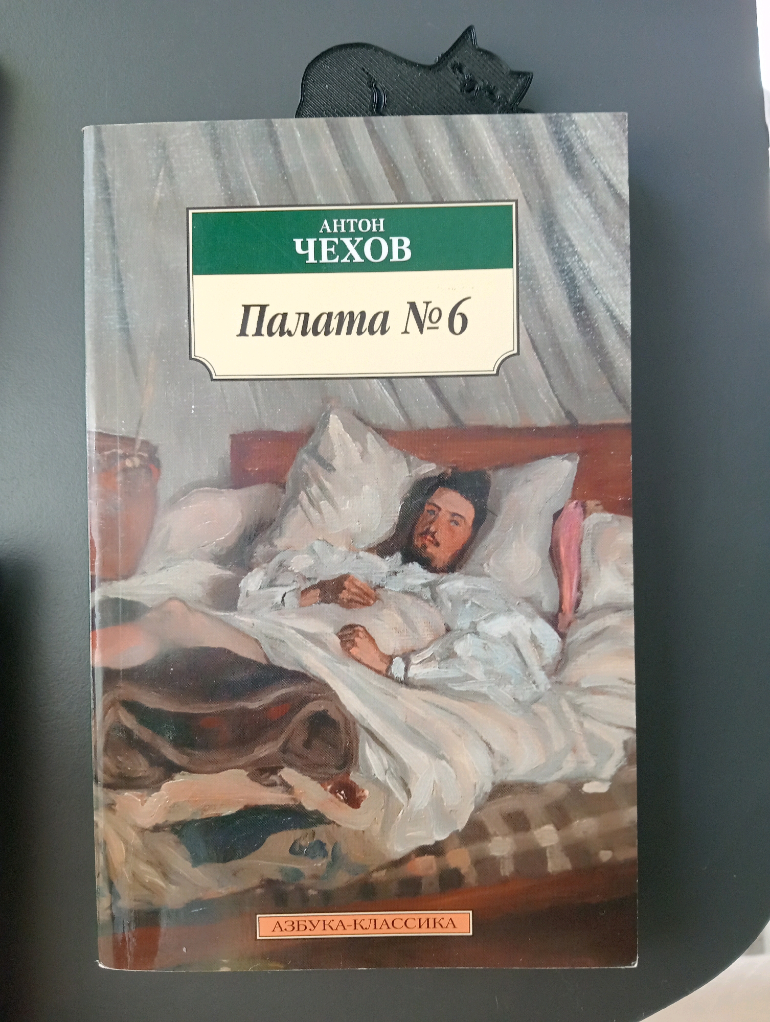 The image shows the cover of a book titled "Палата № 6" (Ward No. 6) by Anton Chekhov. The cover features a painting of a man lying in a bed, surrounded by pillows and blankets. The man has dark hair and a beard and is wearing a white shirt. The painting style is impressionistic. The book has a black cat-shaped bookmark attached to its top edge. The book is resting on a dark gray surface. There is no information in the image that would suggest a location in the city of Silifke, Turkey.