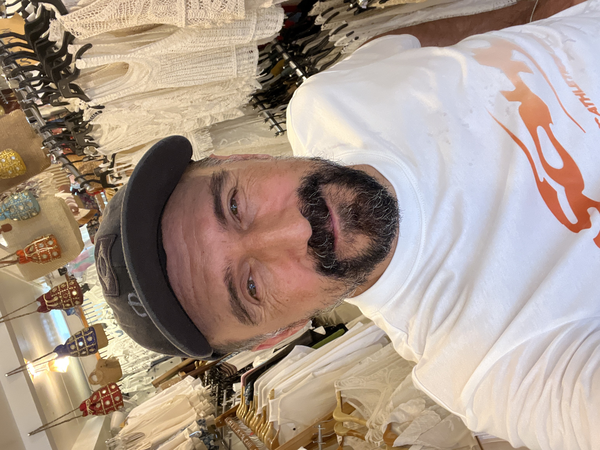 A person wearing a grey cap and a white t-shirt with an orange graphic is posing for a selfie inside a clothing store. The person has a beard and is facing the camera. Racks of clothing, mostly white lace garments, are visible in the background. Small ornamental items hanging from the ceiling are also visible. The location is Baan Plai Laem, Thailand.