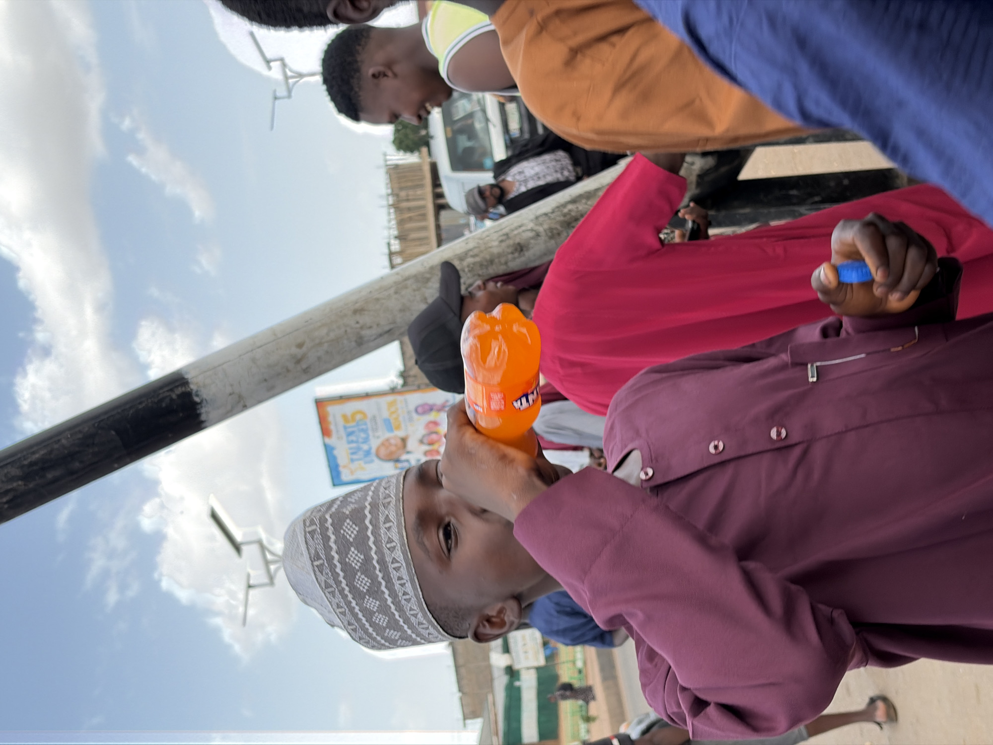 A young man in a grey and white patterned kufi and a maroon garment is drinking from an orange Fanta bottle. He is surrounded by other people, one wearing an orange shirt and another a red garment. A utility pole with black and white markings is visible in the foreground. In the background, a building and a sign with the words "TALENT" are seen. The setting appears to be an urban area in Gura Top, Nigeria, based on the subject's clothing.