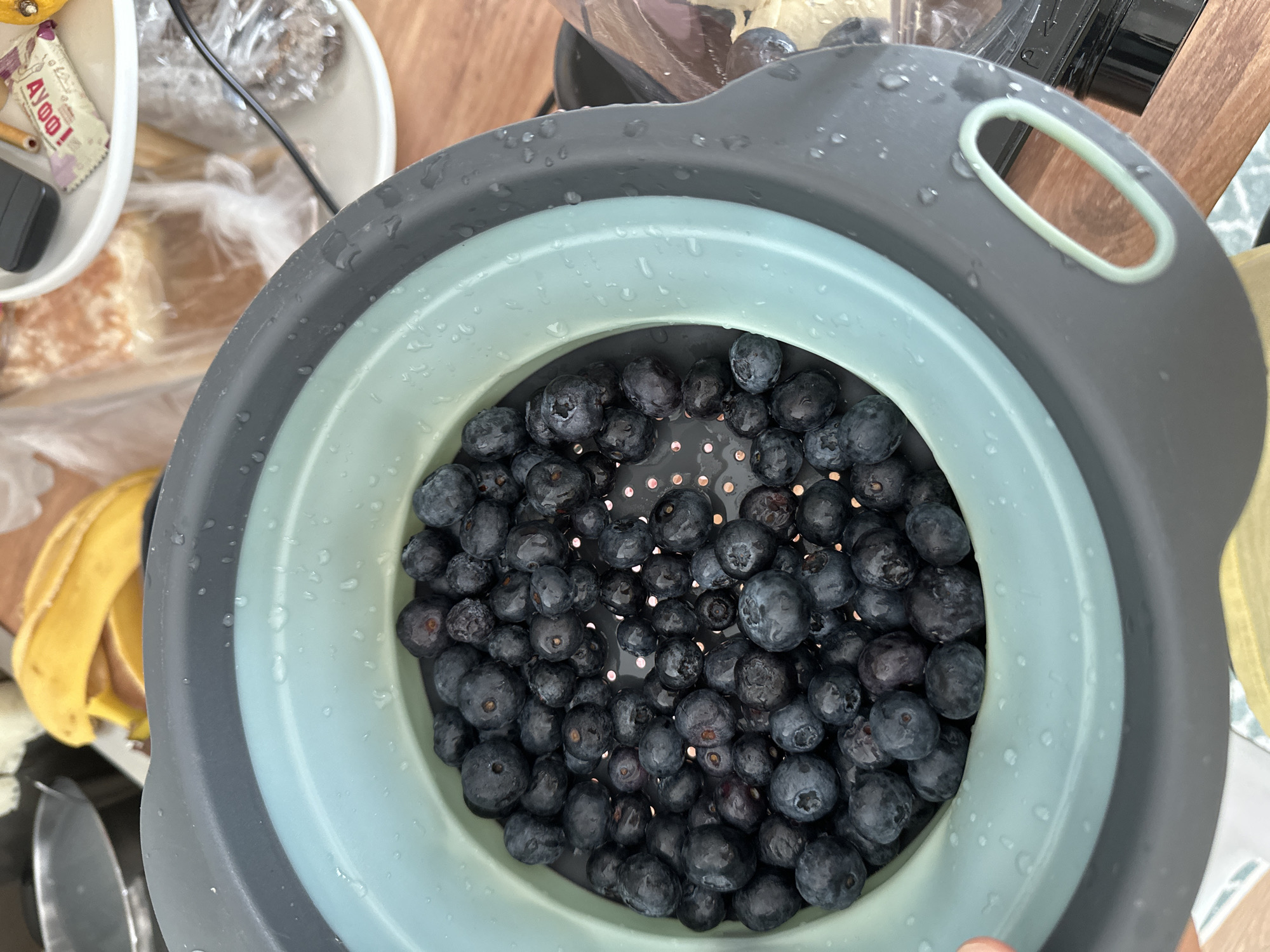 A close-up, high-angle shot shows a collapsible colander filled with fresh blueberries, resting on a wooden surface. The colander is gray and teal, with water droplets visible on its exterior. In the blurred background, a bunch of bananas, a loaf of bread in plastic packaging, and a small wrapped candy are present. The location is Vsevolozhsk, Russia.
