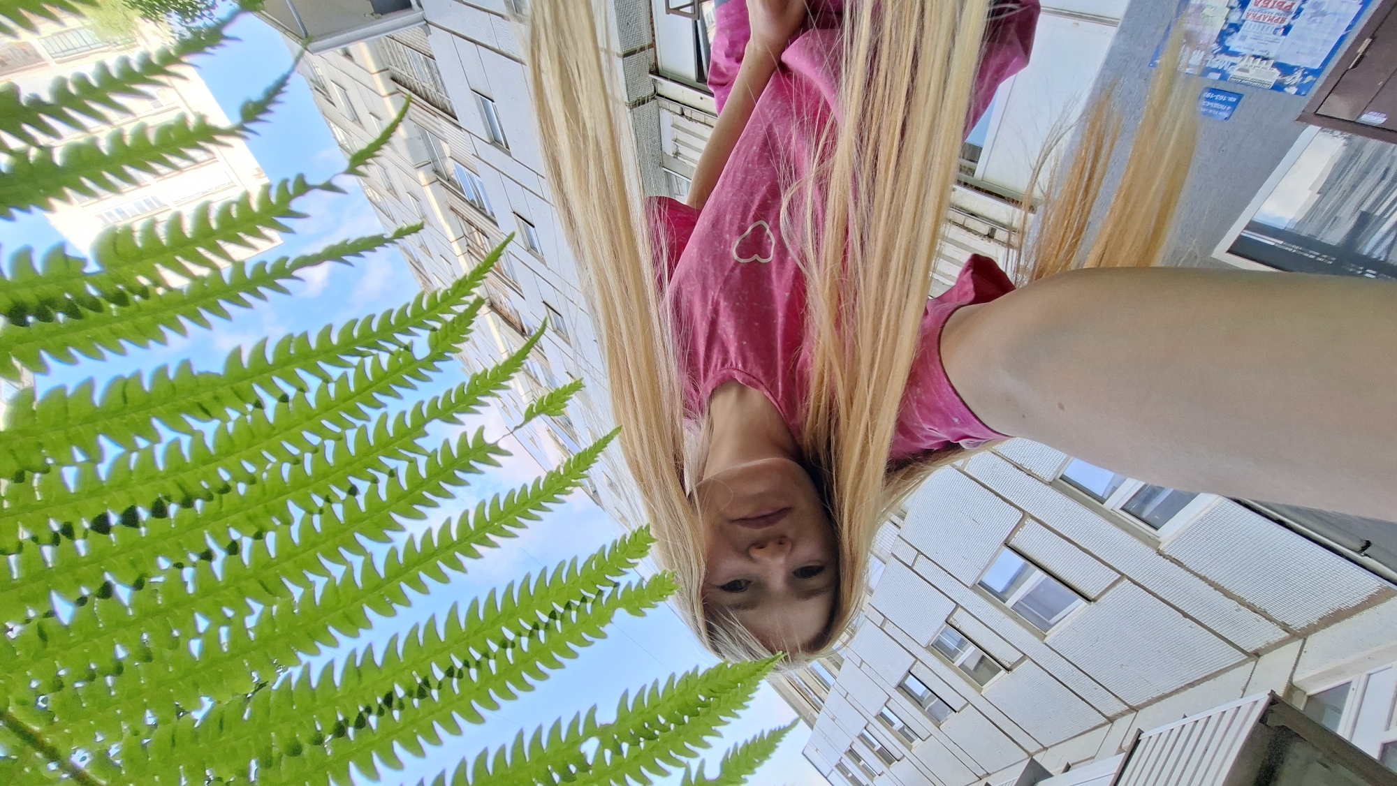 An outdoor, low-angle, wide shot captures a young Caucasian woman with long blonde hair and a pink t-shirt, looking up at the camera. She is partially obscured by the fronds of a green fern plant, which dominates the upper right portion of the frame. Behind her, a multi-story, light-colored apartment building with numerous windows is visible, angled upwards. The sky is blue with some white clouds. The scene is likely a residential area in the city of Chaikovsky, Russia, given the architectural style of the building.