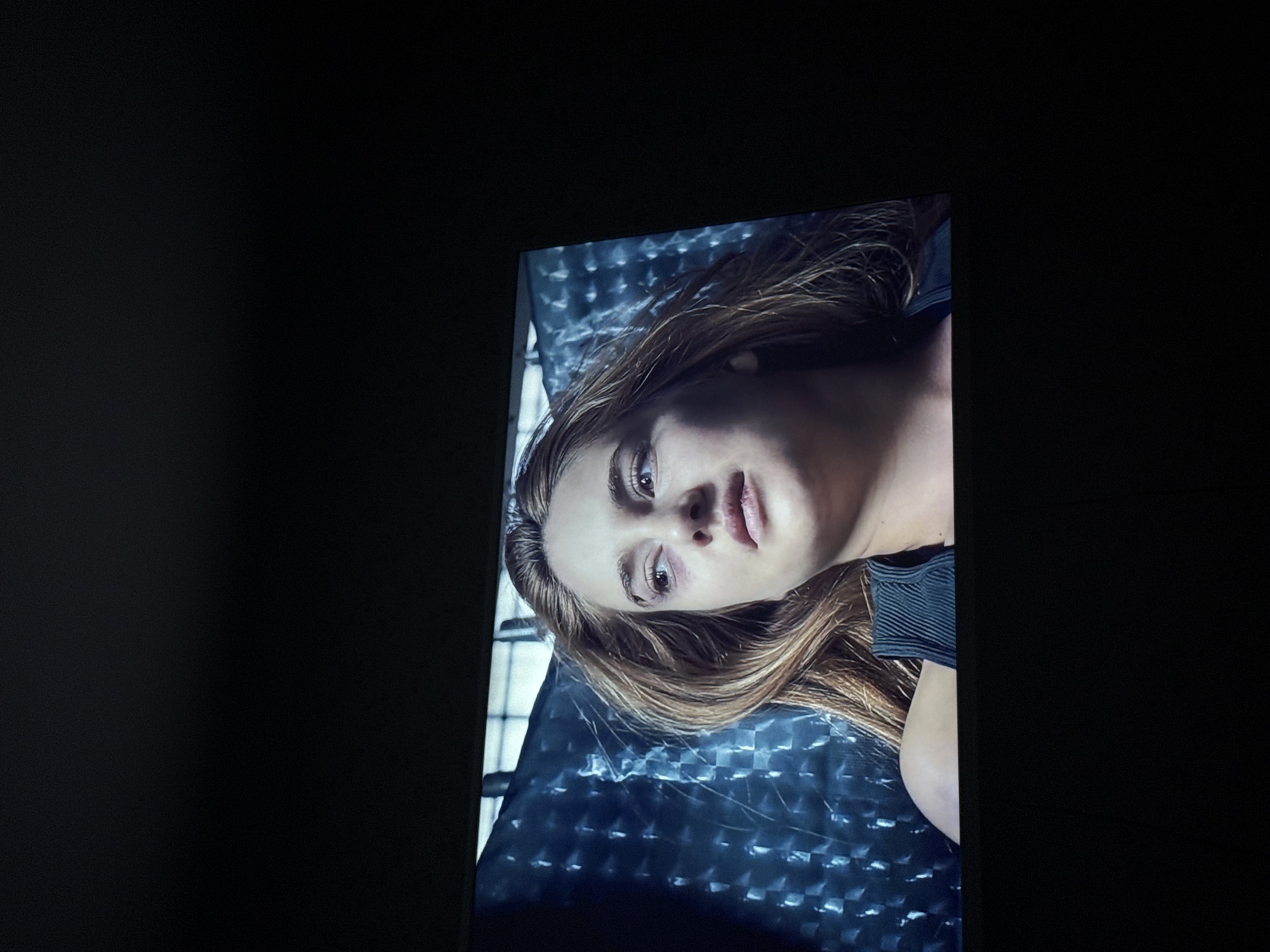 The image depicts a screen displaying a woman lying on a metallic, patterned surface. The woman has light skin, light brown hair, and neutral facial expression. She is wearing a dark, possibly ribbed top. The background behind her is a grid-like metallic surface with a blueish tint. The surrounding area is dark, suggesting the screen is in a dimly lit room. The location is not discernible from the image itself, and there is no evidence to suggest the image was taken in the city of Mytishchi, Russia.