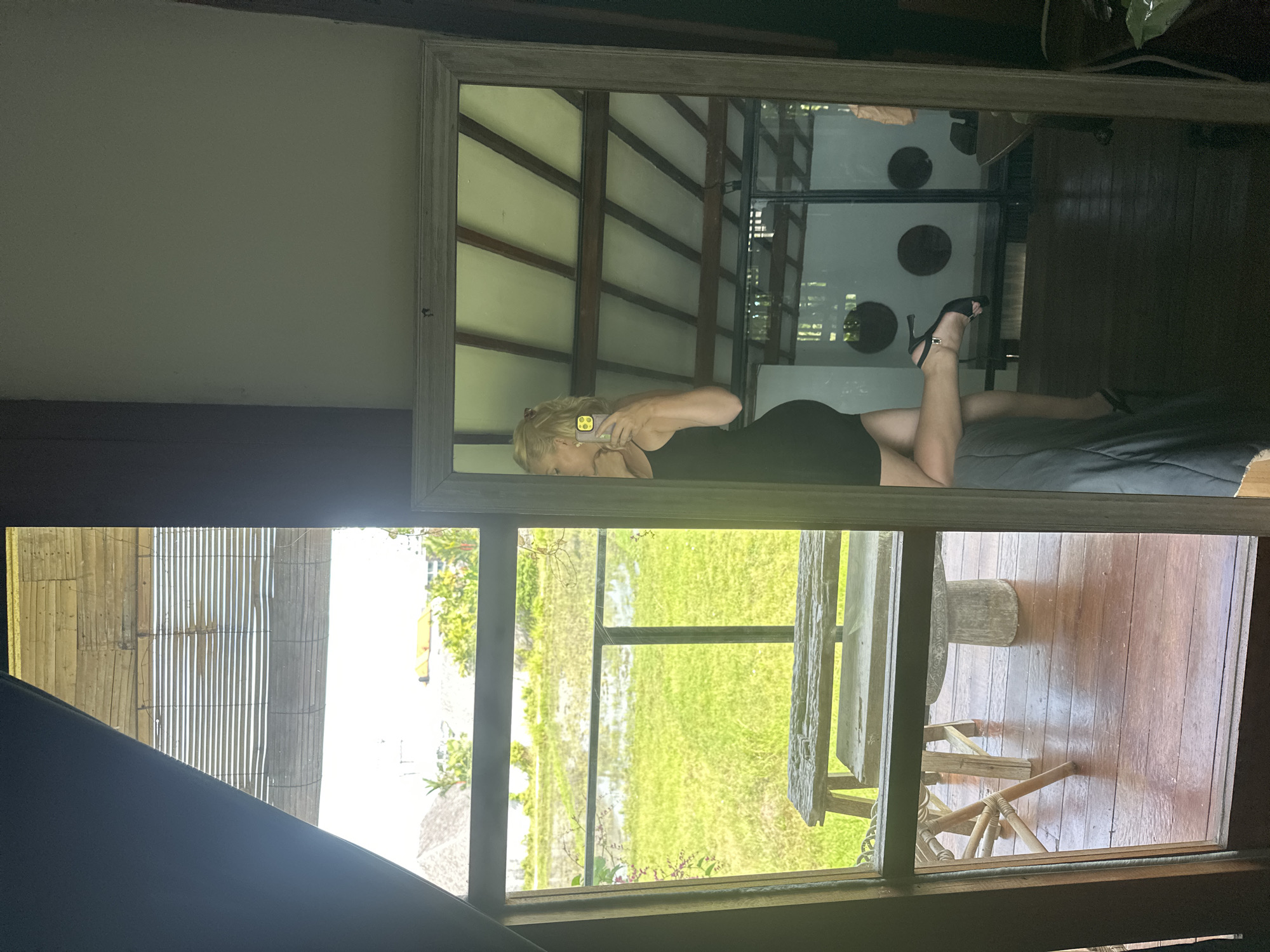 A reflection of a woman is captured in a large rectangular mirror. She is lying on a bed with gray bedding. She wears a black dress and black high heels, and she is holding a cell phone to her face. The mirror reflects a ceiling with exposed beams and a room with dark flooring and circular wall decorations. Outside the window, there is a green landscape with a table and chairs. The image was taken in North Kuta, Indonesia.