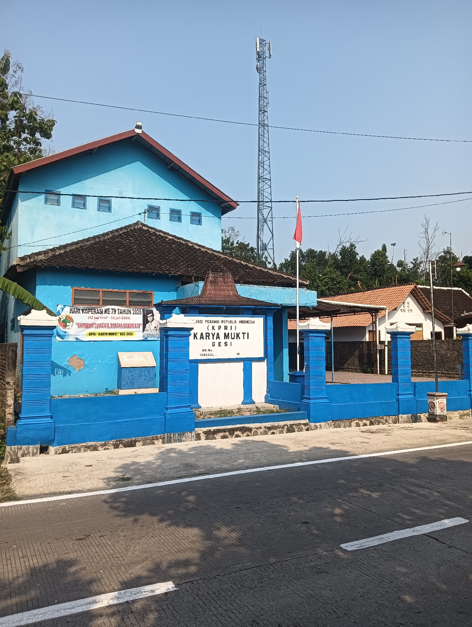 The image shows a bright blue building with a red-tiled roof in Palemrejo, Indonesia. The building features a white sign that reads "YAYASAN PEGAWAI REPUBLIK INDONESIA (KPRI) KARYA MUKTI". The building is surrounded by a blue fence. To the left of the building, a banner with text and images is displayed. A red flag flies from a pole in front of the building. A tall cell tower is visible in the background. The scene is illuminated by sunlight, casting shadows on the road in the foreground. The environment appears to be a rural or semi-urban area with trees and other buildings visible in the distance.