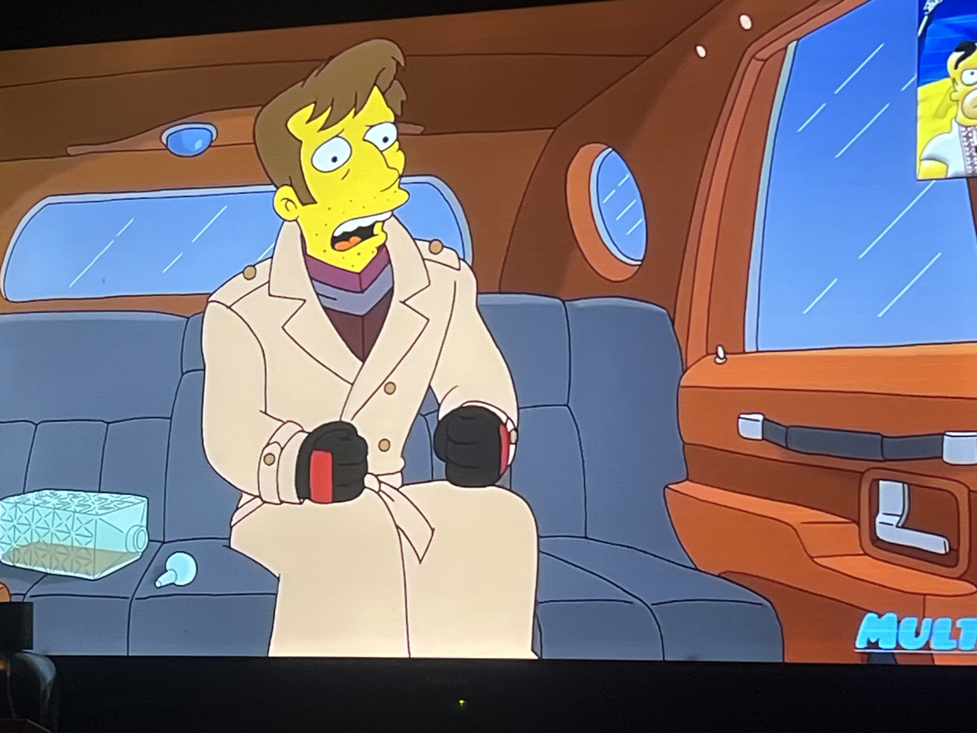 The image depicts a scene from an animated program, likely "The Simpsons," featuring a character with yellow skin and a brown hairstyle, wearing a tan trench coat, a purple scarf, and black gloves with red accents. He is seated in what appears to be the interior of a vehicle, possibly a limousine, with blue upholstered seats and brown wood-paneled interior walls and doors. There is a small, round window on the right side of the vehicle. On the seat next to the character is a translucent blue, geometric container and a small, light blue dropper. A small television screen in the upper right corner shows another animated character, possibly Homer Simpson, wearing a blue shirt with white embroidery. A logo reading "MULT" in blue font is visible in the lower right corner. The background outside the vehicle's windows is a bright blue, suggesting daylight. There is no indication that this scene takes place in Zoriane, Ukraine.