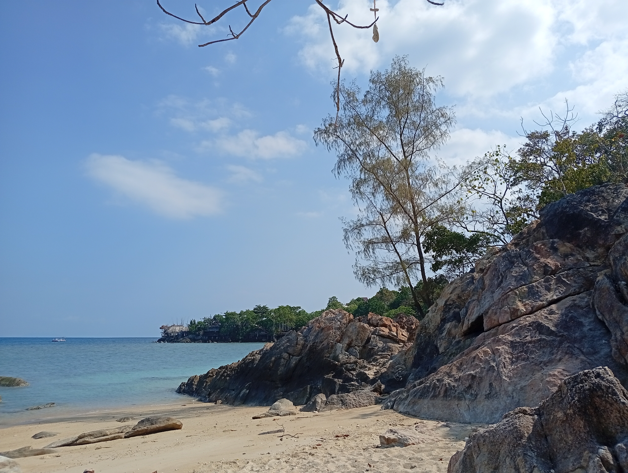 The image depicts a beach scene in Baan Sri Thanu, Thailand. A sandy beach with scattered rocks leads to calm, clear turquoise water. In the distance, a land formation is visible with dense green trees and a building with a wooden structure. A tree with sparse leaves and branches stands on a rocky outcrop, alongside other trees and dense vegetation. The sky is bright blue with scattered white clouds. A few bare branches are visible in the top portion of the frame. The overall scene conveys a sense of tranquility and natural beauty. A small boat can be seen at sea in the distance.