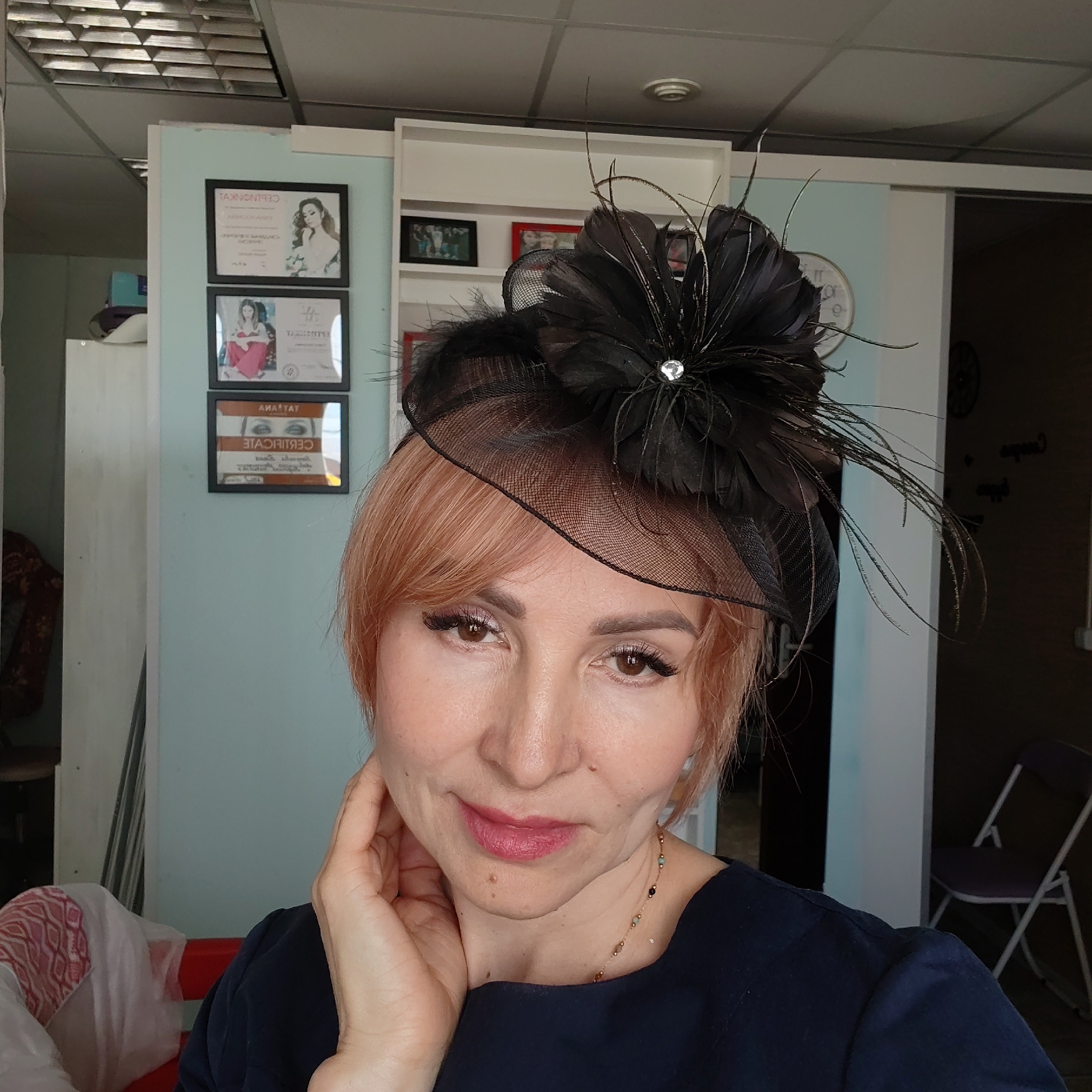 A woman with short, light red hair wears a black fascinator with a net veil, feathers, and a rhinestone embellishment. She is posed with her hand touching her neck. She is wearing a dark blue or black garment and a delicate gold necklace with dark beads.
The background shows a room with light blue walls and white trim. There are framed certificates or portraits hanging on the wall behind her. There is a glimpse of a white cabinet, a folding purple chair, and a partially visible wall with a dark clock.
The location might be Chaikovsky, Russia, given the prompt's mention, but there is no definitive visual evidence in the picture to confirm this. The scene appears to be indoors, possibly in a salon, studio, or home.