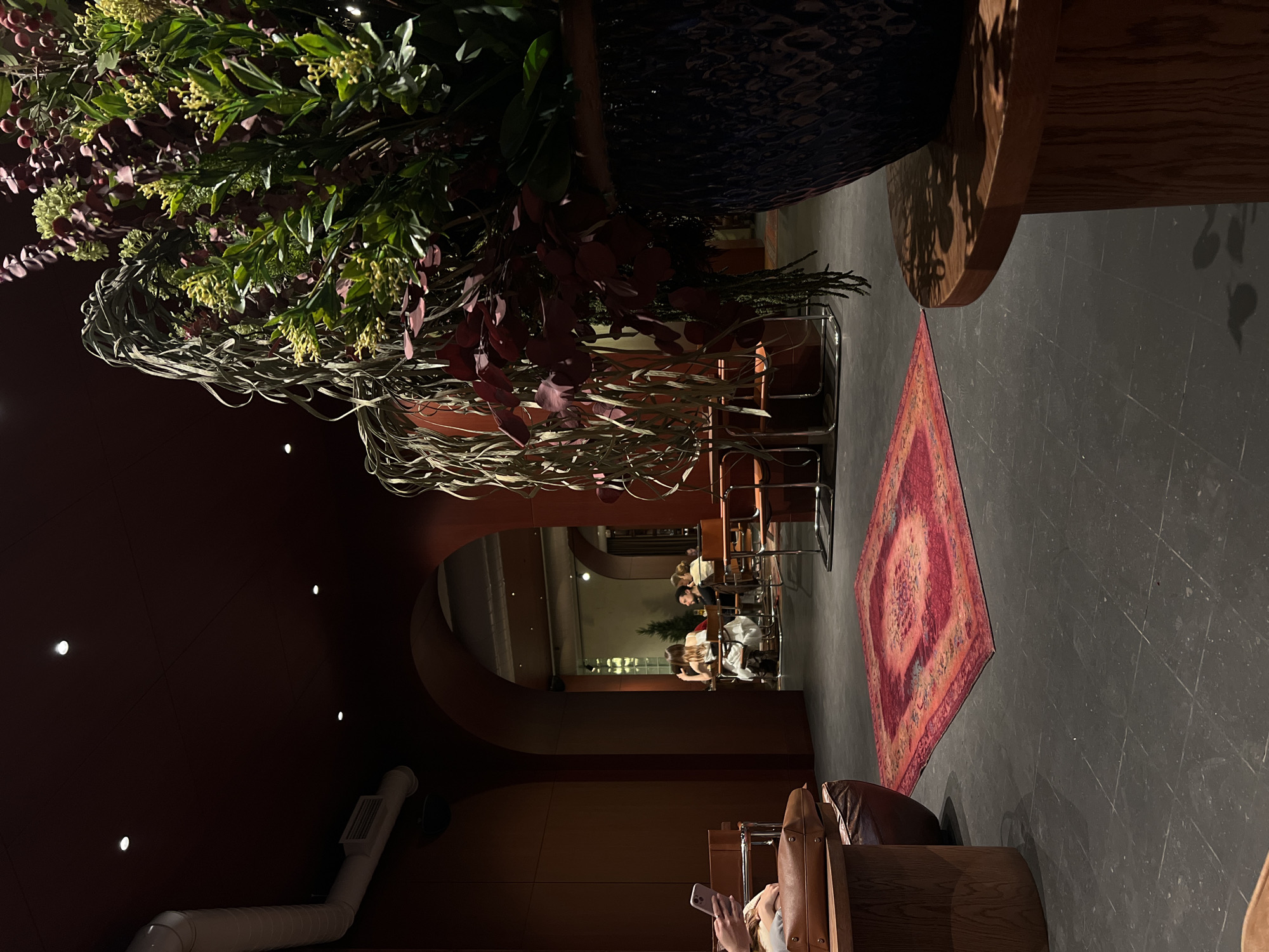 The image depicts the interior of a restaurant, possibly located in Yerevan, Armenia. The decor includes a dark red wall and ceiling on the left, contrasted with lighter wood tones on the right. A large floral arrangement with interwoven branches occupies the upper left portion of the frame. A red patterned rug lies on the dark floor, leading towards a dining area with tables and chairs where people are seated. In the background, there is a glimpse of an outdoor space. In the foreground, a person is seated, using a smartphone. The room is illuminated by recessed lighting in the ceiling.