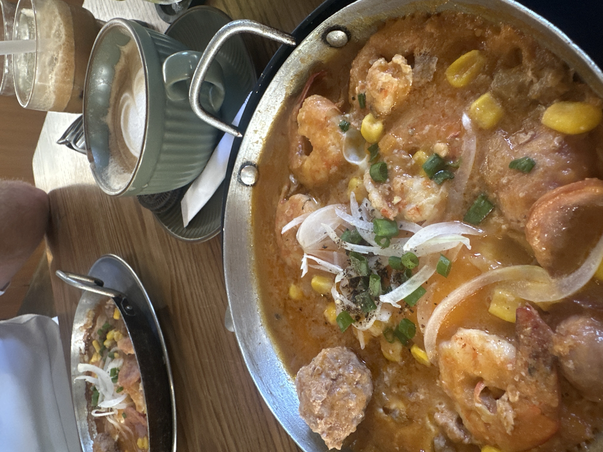 An overhead shot shows food and drinks on a wooden table. A stainless steel pan contains a shrimp and corn dish garnished with green onions and sliced white onions. Beside it, a smaller pan contains a similar dish. Next to the pans is a green ceramic cup of coffee, and a tall glass of iced coffee. An arm and part of a white shirt are visible on the left side of the frame. The location is likely Bac Nha Trang Ward, Vietnam.