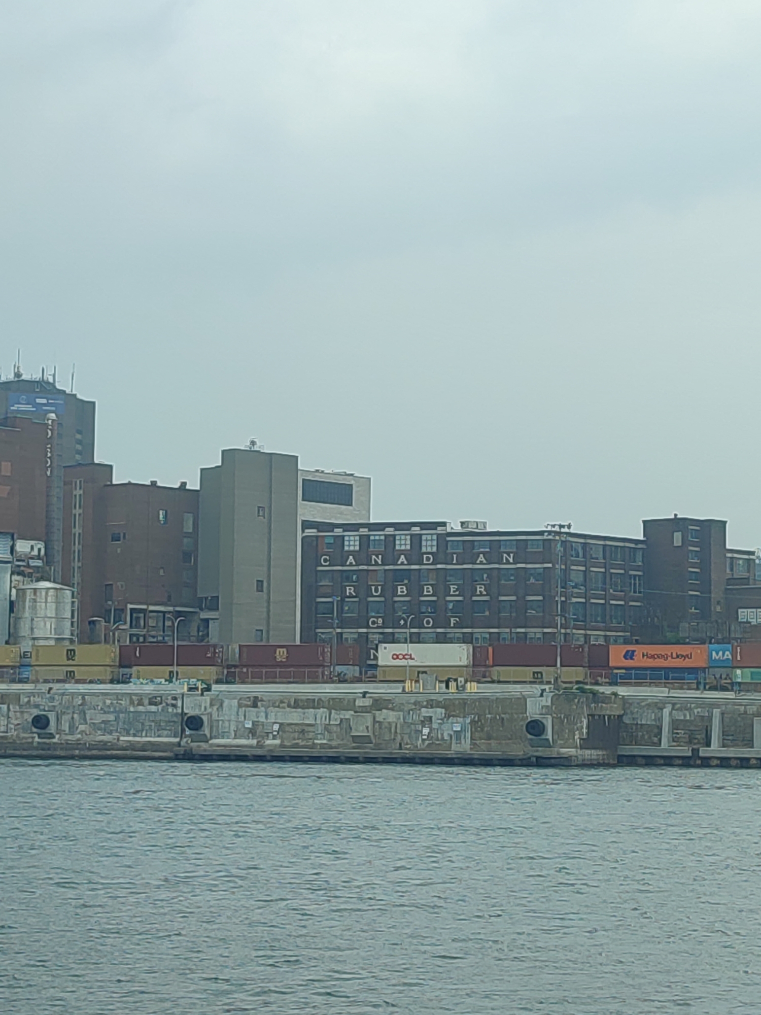 This is a photograph of the Old Port of Montreal, Quebec, Canada, on a hazy day. In the mid-ground, a large brick building with "CANADIAN RUBBER" inscribed on its facade dominates the scene. Several other industrial buildings, some taller and modern, are visible behind and to the left of the brick structure. In the foreground, the murky waters of the St. Lawrence River are visible, lapping against a concrete retaining wall. Several shipping containers are stacked on the waterfront. The sky is overcast and a pale blue-gray.