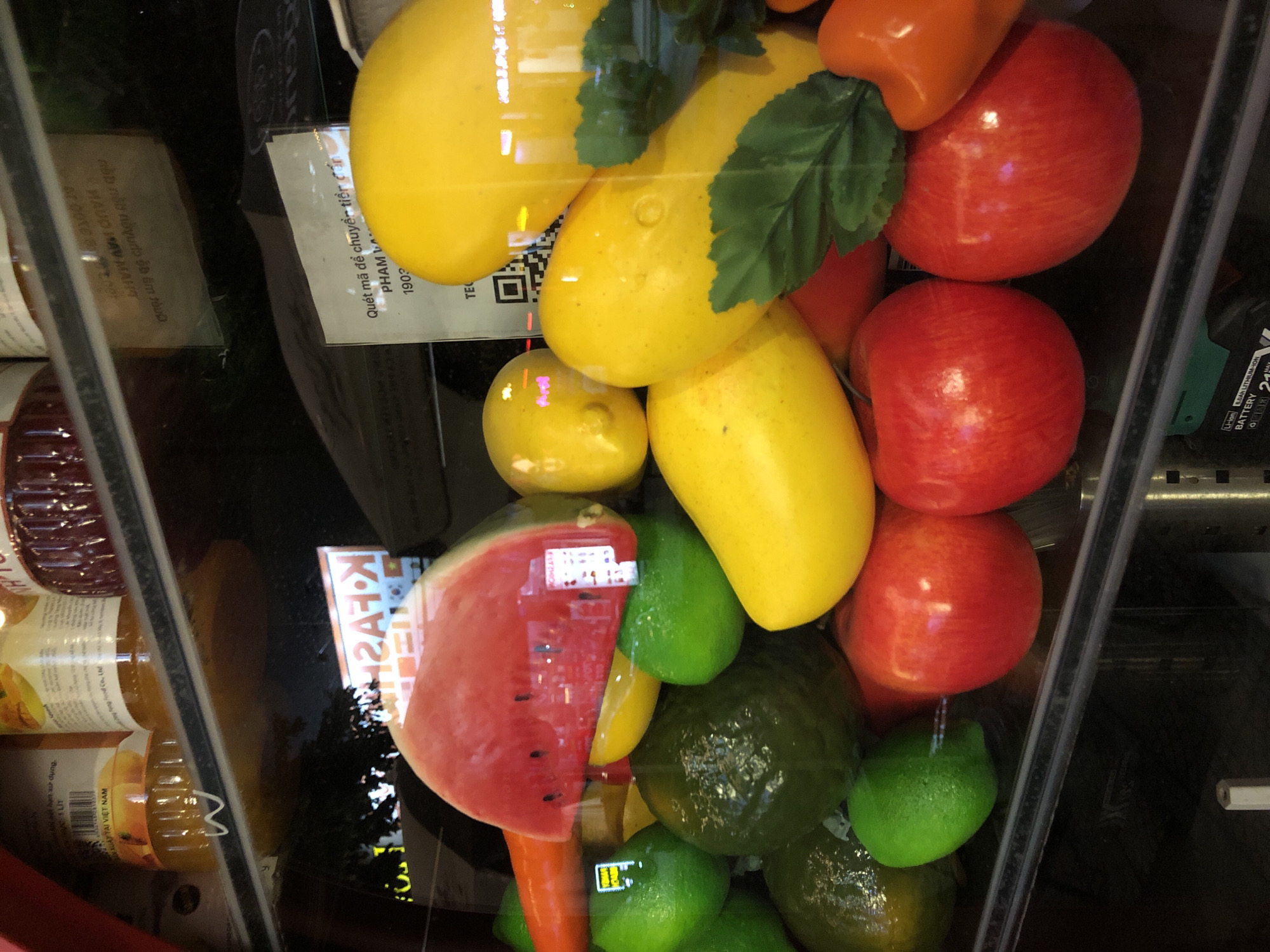 This is a medium shot, slightly angled, indoor photograph of a display of artificial fruit within a glass-fronted refrigerator or cooler. The focus is on a colorful assortment of fruits, including yellow mangoes, red apples, a slice of watermelon, green limes or avocados, and a single orange fruit at the top. The fruits appear to be of various sizes and are arranged in a somewhat chaotic but visually appealing manner.
Behind the fruits, a reflective surface, likely the glass of the cooler, shows some blurred reflections. There are also labels and text visible on some of the items within the display and on the cooler itself. One prominent label appears to be a QR code with Vietnamese text above it, which translates to "Scan the code to transfer money to PHAM A." Other visible text includes what seems to be product information, "MADE IN VIETNAM," and battery-related text.
The lighting suggests an indoor setting, possibly a market, grocery store, or a food service establishment. There are no people visible in the frame. The overall impression is a vibrant and plentiful display of fruit, designed to attract attention, with subtle hints of a commercial environment. The image is sharp and well-lit, allowing for clear observation of the textures and colors of the artificial fruits. The date and location context suggest this might be a snapshot taken while shopping or visiting a store in Da Nang, Vietnam.