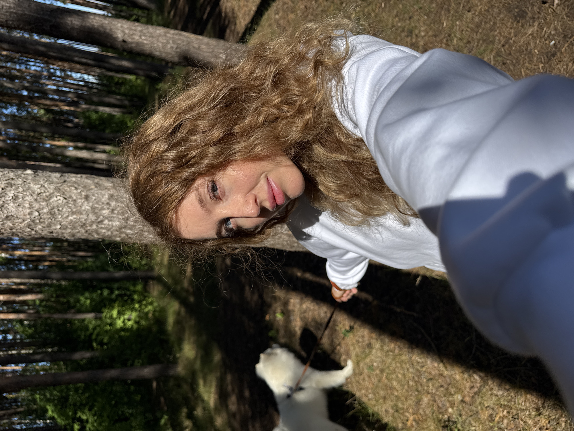 In a sun-dappled forest setting, likely in Chaikovsky, Russia, a young woman with wavy, reddish-blonde hair is taking a selfie. She is wearing a white hooded sweatshirt and has a light complexion with rosy cheeks and pink lips. Her blue eyes are looking towards the camera. To her left and slightly behind her, a fluffy white dog, possibly a Samoyed, is walking on a leash. The dog is out of focus. The background consists of tall, slender pine trees and dense green foliage, indicating a natural, wooded environment. The ground is covered with dry grass and pine needles, and shadows from the trees create a dappled effect on the ground. The woman is holding a leash attached to the dog, suggesting they are on a walk.