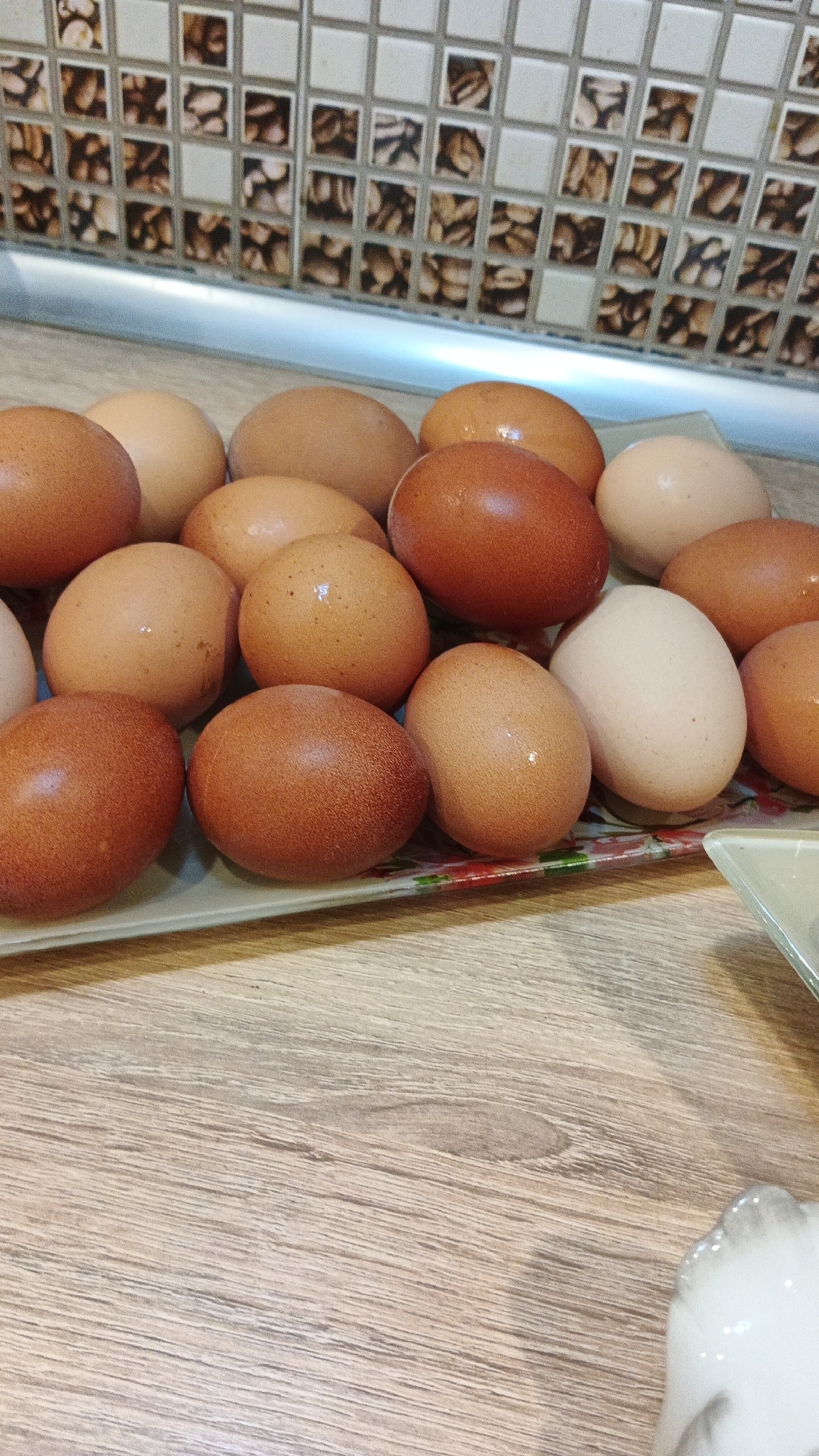 A plate of farm fresh brown and white eggs sits on a wooden countertop. The eggs are arranged in a somewhat haphazard manner, with some overlapping. The lighting suggests it is daytime, and the scene appears to be indoors, likely a kitchen. The background features a tiled backsplash with a mosaic pattern. No people are visible in the image. The image does not contain any discernible text. The overall impression is one of a simple, domestic scene.