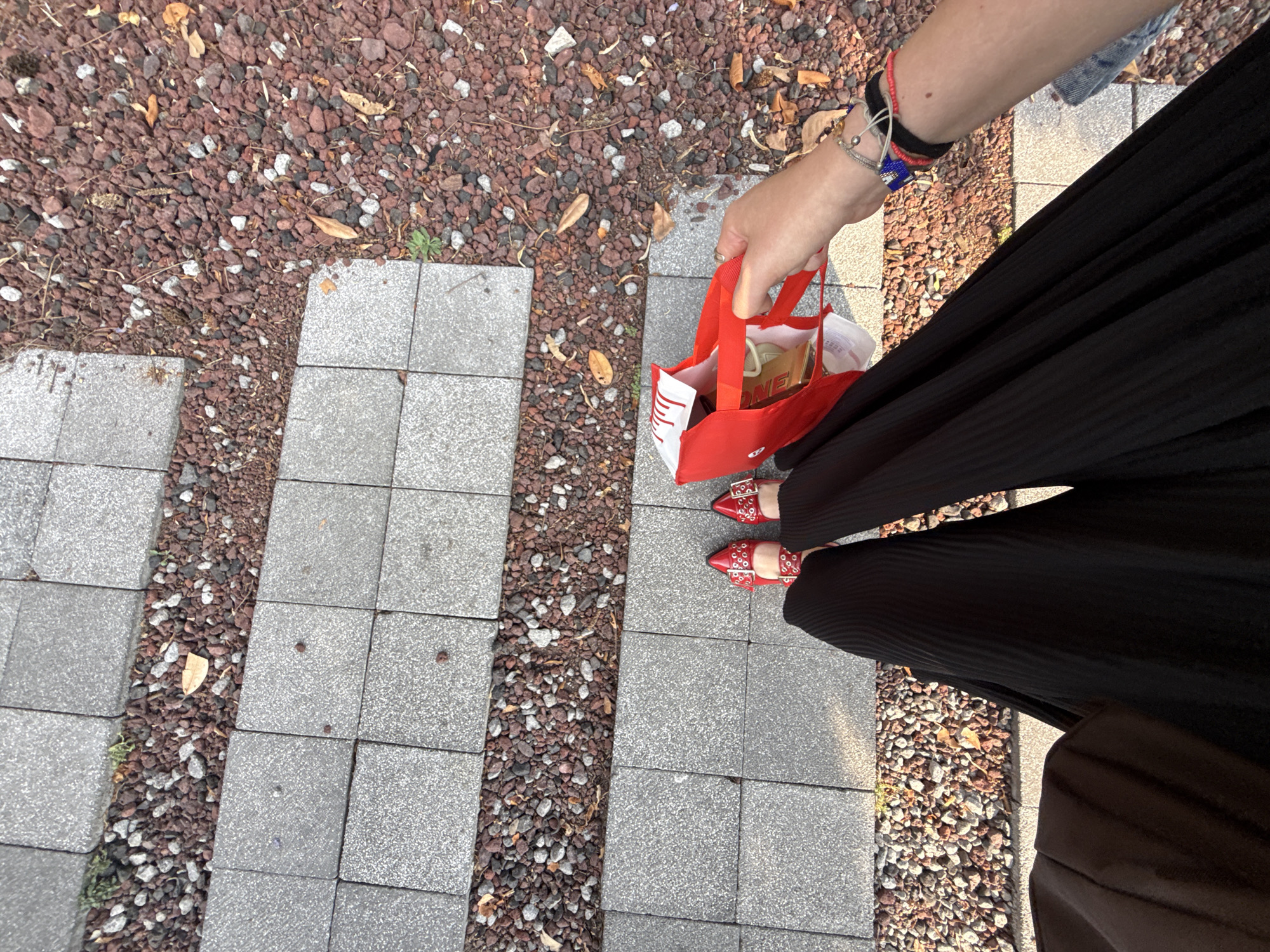 The image captures an overhead view of a person's lower body and arm, positioned on an outdoor path. The path features sections of light gray square concrete pavers alternating with strips of reddish-brown gravel, interspersed with dried leaves and small debris.
The person is wearing wide-leg black pleated pants or a long skirt, paired with eye-catching red flat shoes adorned with silver buckles and studs. From the upper right, an arm extends into the frame, adorned with multiple bracelets including red, black, silver, and blue elements. The hand holds a small, bright red shopping bag.
Inside the red bag, several items are visible, including a golden-yellow package with the word "ONE" clearly discernible. Other white packaging or labels with obscured text and a partial barcode are also present. Some white text is partially visible on the side of the red bag.
The scene is brightly lit by natural sunlight, suggesting daytime with clear weather. The setting appears to be an urban or park pathway, consistent with general outdoor public spaces in Mexico City, Mexico, as per the provided context. The person appears to be standing or walking slowly, holding the bag.