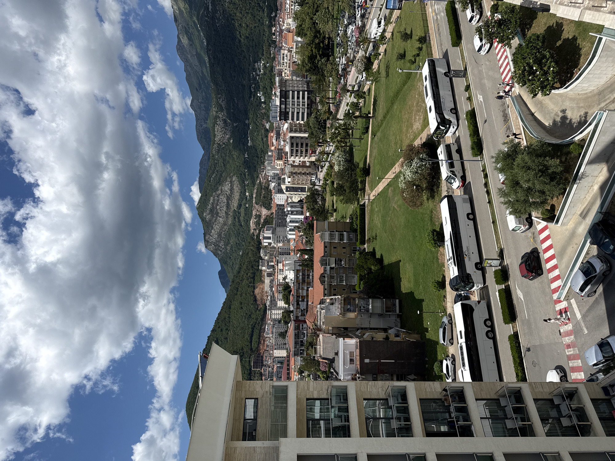 This is a high-angle, medium shot of a street scene in Budva, Montenegro, on a sunny day with scattered clouds. In the foreground, a modern apartment building with balconies is visible on the left. Across the street, several large white tour buses are parked, and a white van is also present. Pedestrians are crossing a red and white striped crosswalk. Beyond the buses, there is a well-maintained grassy park with palm trees and other foliage. Further in the distance, a dense urban area with many buildings, most with red-tiled roofs, stretches towards a backdrop of lush green mountains. The city is built on the slopes of these mountains, which are a prominent feature of the background. The sky is a vibrant blue with dramatic white and grey clouds.