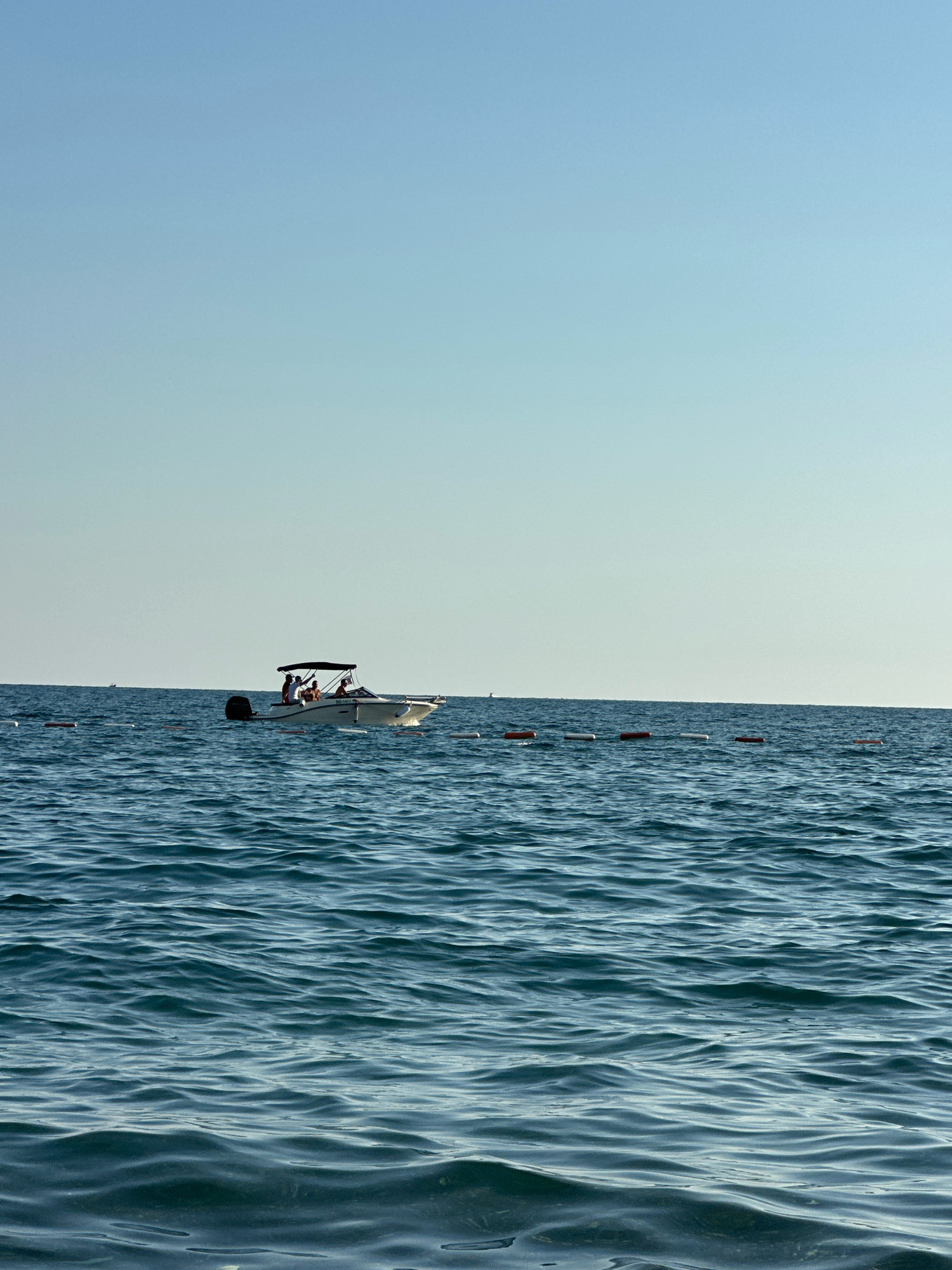 A motorboat with several people on board is sailing on the sea under a clear blue sky. The boat is white with a dark canopy and an outboard motor. The sea is a deep blue with small waves. There are buoys in the water, marking a boundary. The scene is located off the coast of Boreti, Montenegro.
