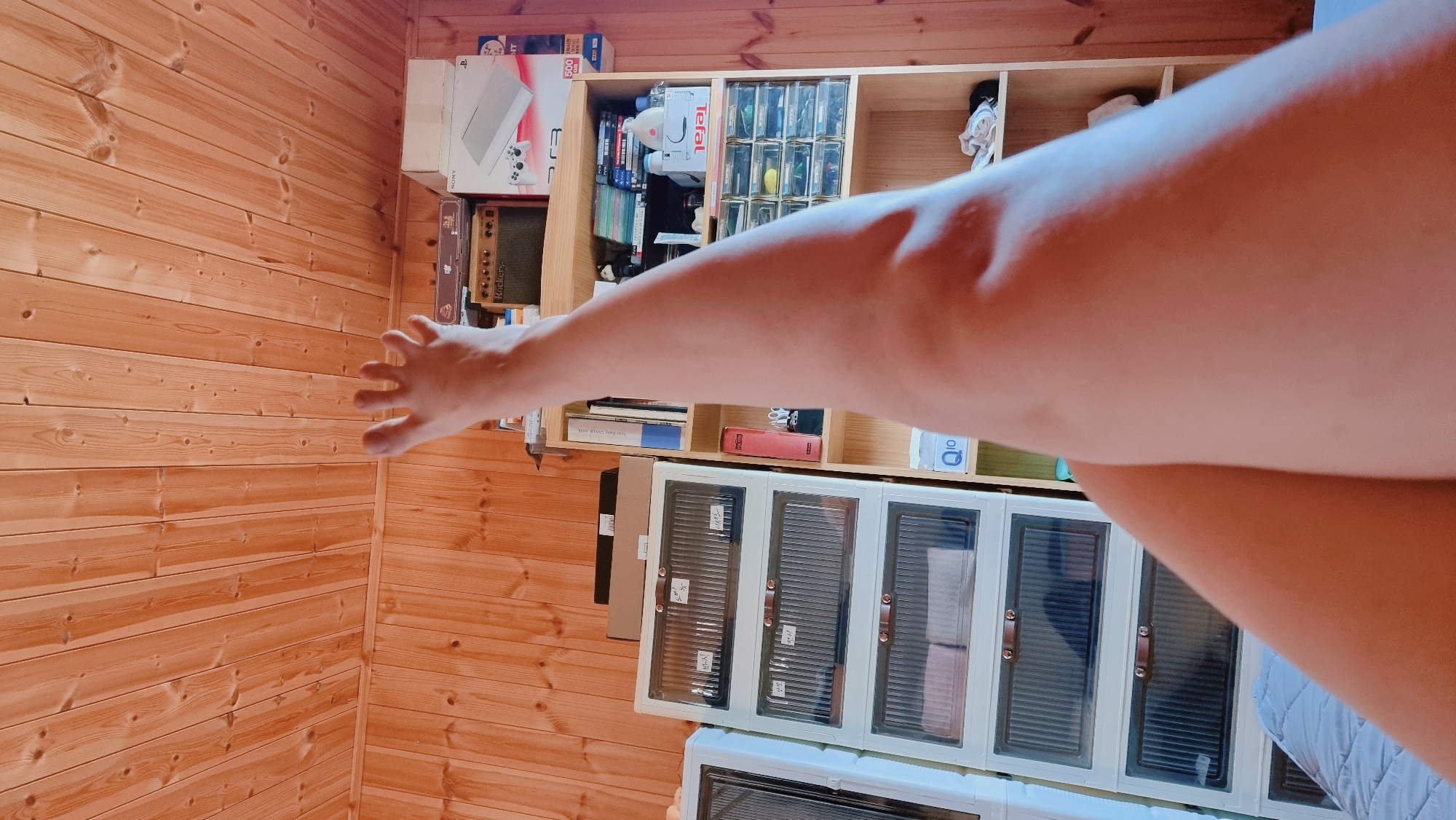 A photograph taken indoors in Dunnae-myeon, South Korea, shows a person's arm and hand extended toward a wooden wall. The person is likely holding a camera to take the photo. In the background is a wooden shelf containing various items, including boxes, books, and storage containers with clear doors. The floor and wall are made of wood. A blue quilted blanket is partially visible in the lower right corner.