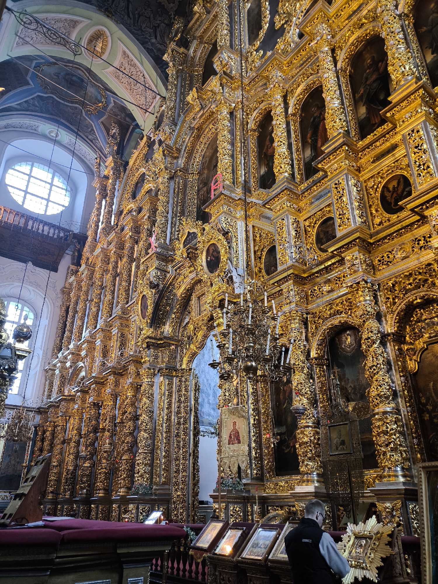 The image captures the interior of a richly decorated Orthodox church, likely the Cathedral of the Assumption in Smolensk, Russia. The architecture is predominantly gold-toned, featuring intricate ornamentation and numerous religious icons within the walls and structures. A large, arched window allows natural light to filter into the space, highlighting the ornate details. In the foreground, a man is looking at, or possibly adjusting, a framed icon or image on display. There is a red bench or seating area with small lights or candles, possibly indicating an area for prayer or reflection. The environment exudes an aura of religious reverence, characterized by its grandeur and meticulous detail.