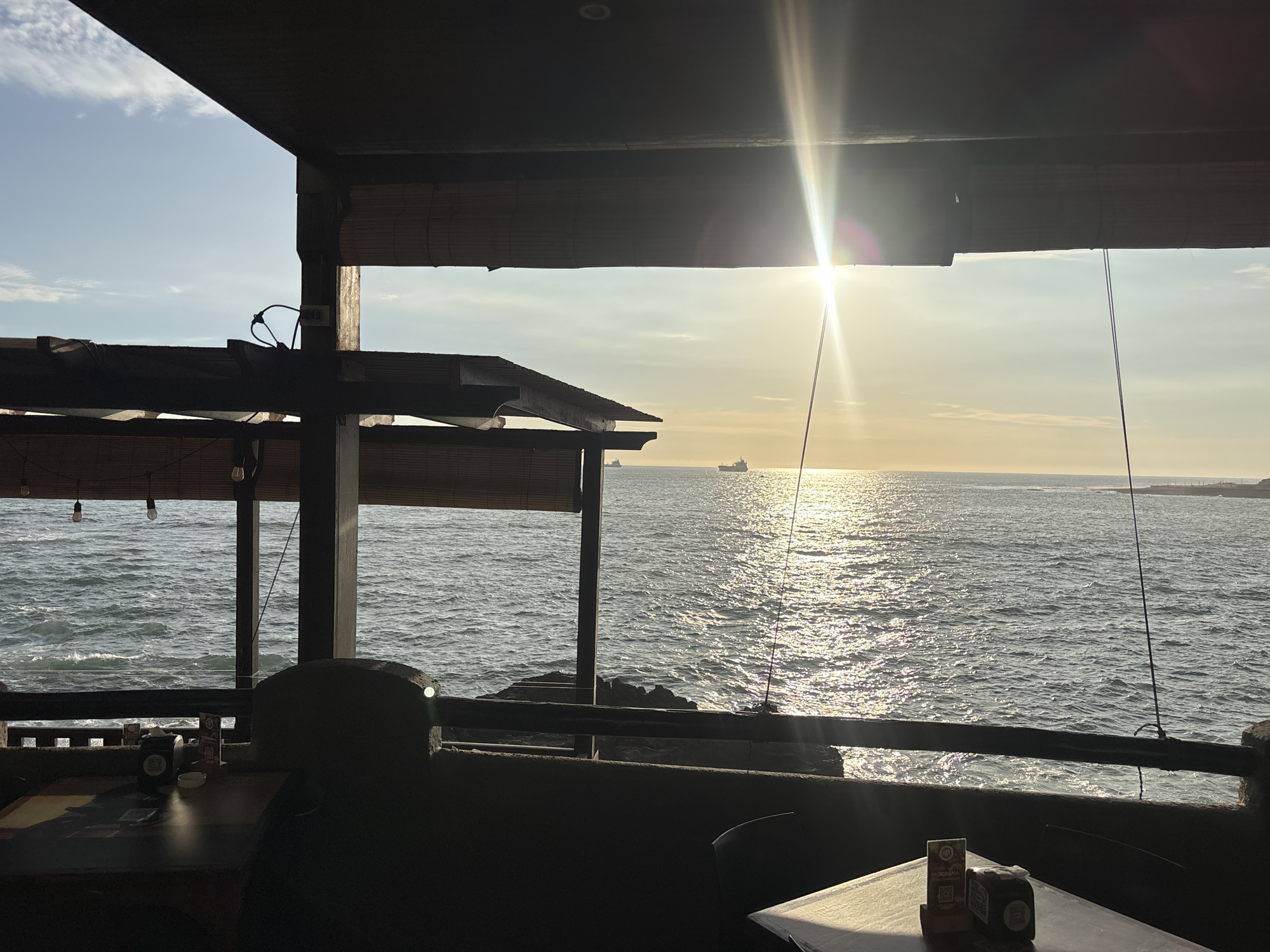 The image presents a scenic view of the ocean from a dimly lit, wooden-structured interior, likely a coastal restaurant or cafe in Arica, Chile. The perspective looks outwards, framed by the building's architecture.
In the foreground, the establishment's interior is largely in shadow, showcasing dark wooden beams, a ceiling, and parts of rolled-up bamboo blinds. Several tables and chairs are visible, also mostly darkened. On the table closest to the viewer, there's a napkin dispenser, a small white bowl, and a cardboard stand. Hanging light bulbs are suspended from the overhead beams. A low, dark railing forms a barrier along the edge, separating the seating area from the open sea.
The midground and background are dominated by the expansive ocean. The water surface is sparkling intensely, indicating strong sunlight reflecting off the waves, creating a prominent sunburst effect in the upper right. The sun appears low on the horizon, suggesting a late afternoon or early evening setting. In the distance, several large ships or cargo vessels are visible against the horizon. The sky is a pale blue with scattered, wispy clouds. Hints of a rocky coastline can be seen near the water's edge on the left.
Notable details include the stark contrast between the shadowed interior and the brightly lit exterior. The intense sun glare is a key feature, making the ocean shimmer. The scene evokes a tranquil and picturesque atmosphere. On the cardboard stand on the table, the word "PROGRAMA" is clearly visible, along with other smaller, illegible text.