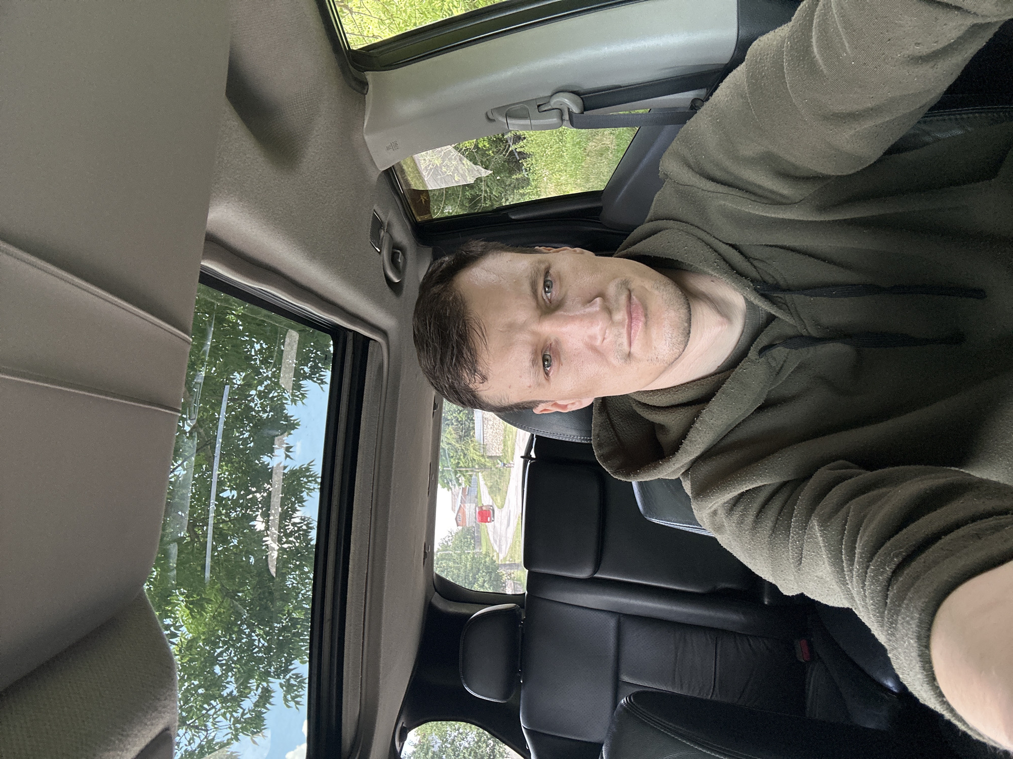 A man is taking a selfie inside a car. The man has short brown hair and is wearing a dark green hooded sweatshirt. The car has black leather seats visible in the background, as well as a sunroof which is open and shows trees and sky. The car appears to be parked on the side of a road with trees and greenery on either side. In the distance, there is a red vehicle on the road and several houses. The setting appears to be rural, possibly in Makiv, Ukraine, given the information provided about the location.