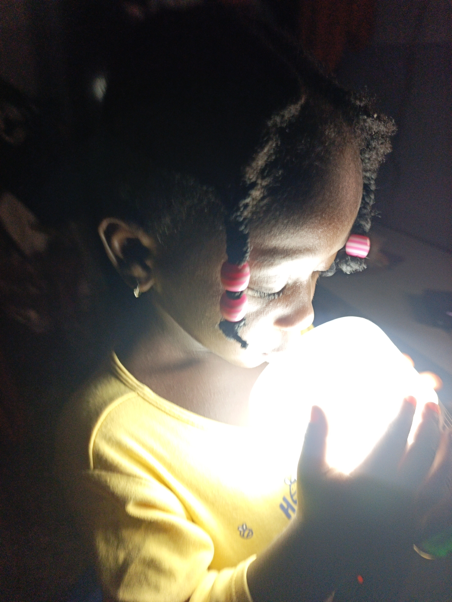 In an indoor, dimly lit environment, a young Black girl is captured in a close-up shot, holding a luminous, spherical object that emits a bright white light. The girl's face is partially obscured as she gazes at the object. She has dark hair styled in braids adorned with pink and white striped beads. She is wearing a yellow long-sleeved shirt, which appears to have a faint graphic print on it. Her right ear, with a small silver earring, is visible. Her hands are cupped around the light source, with her fingers illuminated by its glow. The background is dark and indistinct, providing no specific environmental context for the location within the city of Unknown, Unknown.