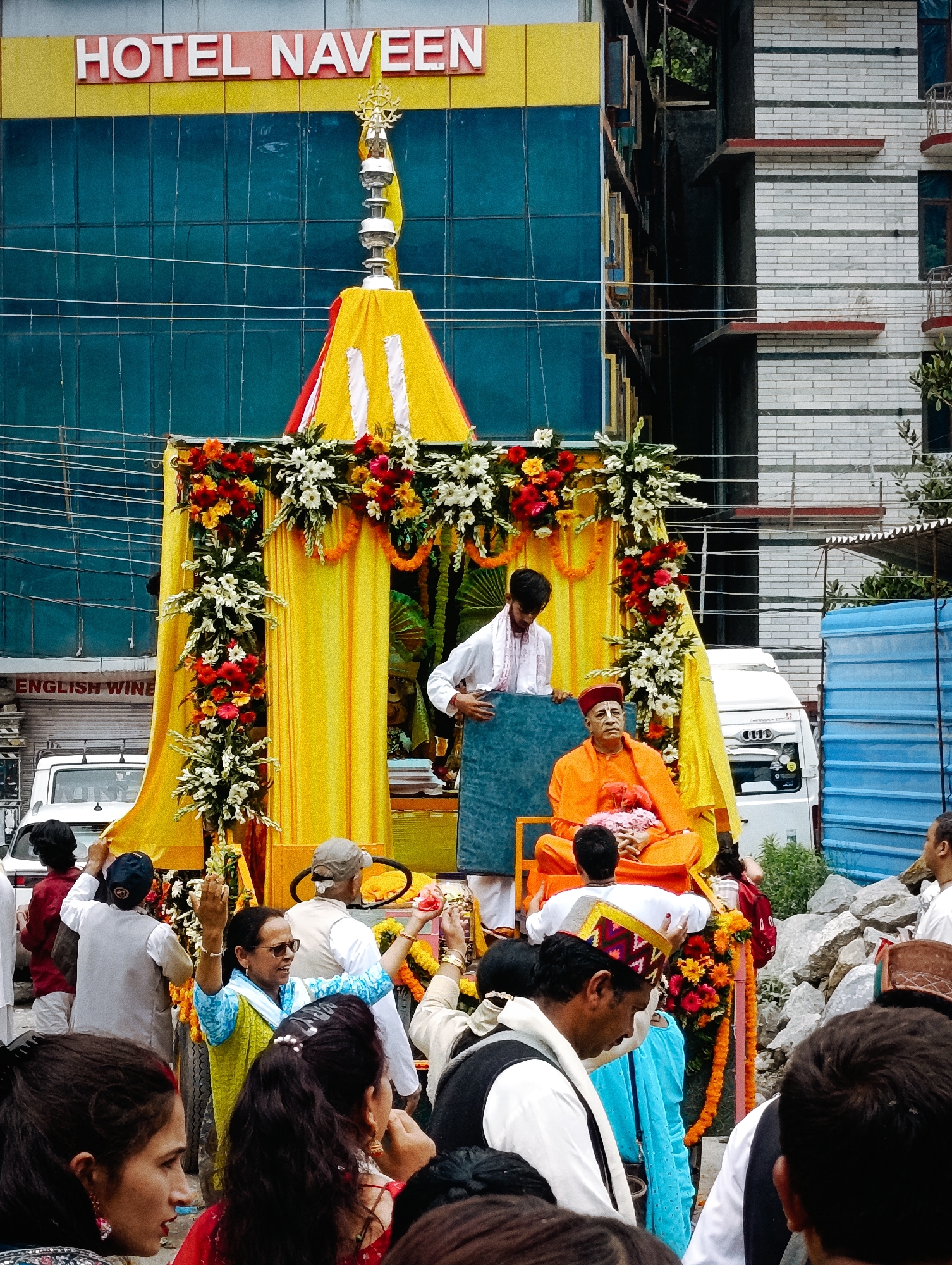 The image depicts a procession in Manali, India, likely for a religious or cultural event. In the background, the "HOTEL NAVEEN" sign is visible above a building with blue glass windows. In the foreground, a decorated float or stage is set up, draped with yellow fabric and adorned with colorful flowers, including red, orange, and white varieties.
Seated on the float is an older man dressed in bright orange robes, wearing a red cap, and holding pink flowers. He appears to be the central figure of the procession. To his left, a younger man in a white shirt and scarf stands, holding a large blue rectangular object.
Several people are gathered around the float, some watching the proceedings, others participating. Among them are women in vibrant clothing, including a woman in a blue and green outfit with her hands raised, holding a red flower. Men in traditional Indian attire, such as kurtas and vests, are also visible. A white van is parked behind the float, and a white car is partially visible on the left side of the frame. The ground appears to be a street, with loose stones scattered in the lower right portion of the image. The overall scene suggests a festive and lively atmosphere.