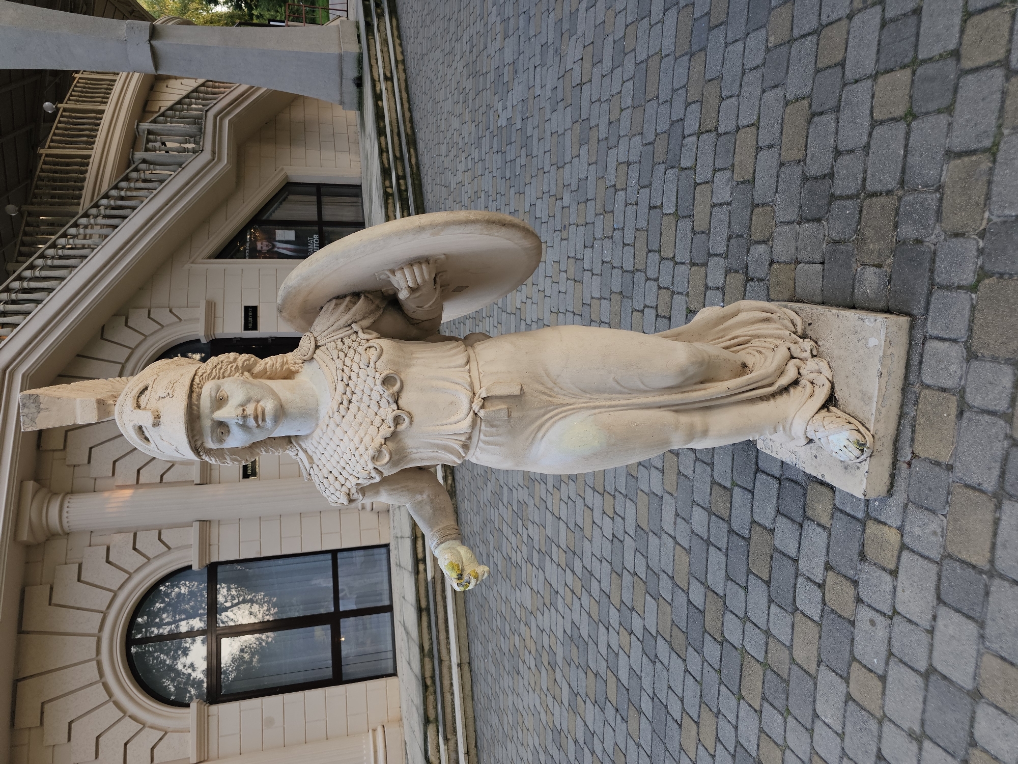 A life-sized, light-colored statue of a woman in ancient armor stands on a paved courtyard in front of a building. The statue depicts a female warrior wearing a helmet, a segmented collar, and a draped gown. She holds a round shield on her left arm and her right hand is open, with what appears to be yellow and white paint on her fingers. The building behind her is light-colored, with arched windows and decorative columns. A large staircase with balusters is visible on the right side of the building. The courtyard is paved with interlocking grey and tan rectangular stones. The statue is situated in front of the "Medpunkt" (medical facility) at the park "Lyubovny Sad" (Love Garden) in Cherkessk, Russia.