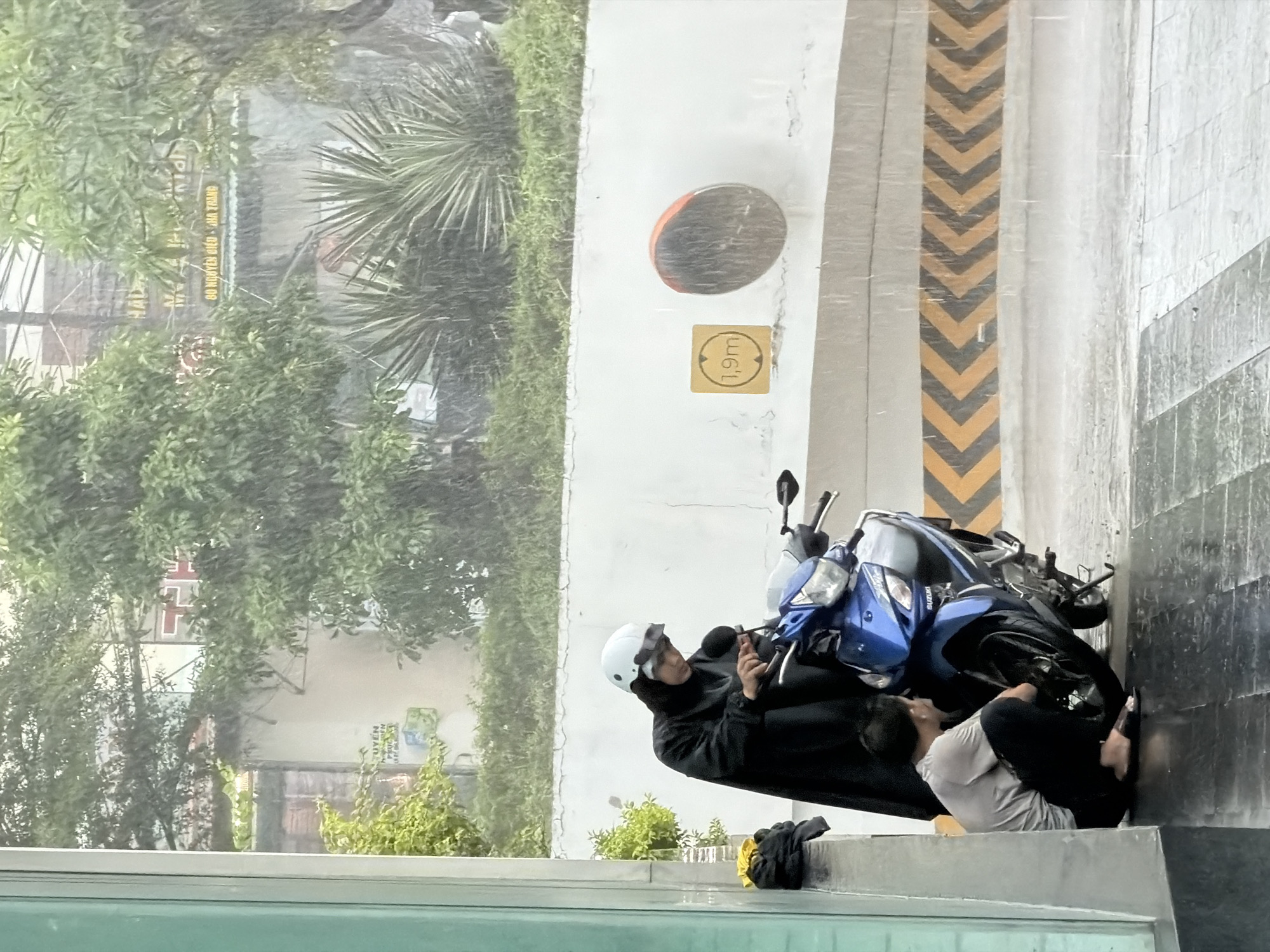The image, taken during heavy rain, shows a scene likely in Nha Trang Ward, Vietnam. The setting appears to be an outdoor parking area near a building. A blue motorcycle is parked at an angle to a wall marked with yellow and black hazard stripes. Two people are sheltering near the motorcycle. One person, wearing a white helmet and dark jacket, is standing and looking at a mobile device. The other person, wearing a light-colored shirt and dark pants, is crouching near the motorcycle and appears to be barefoot. The background includes foliage, buildings, and a sign, possibly indicating the location as "60 NGUYEN GIAC NHA TRANG". The rain is heavy, obscuring details and creating a blurry effect.