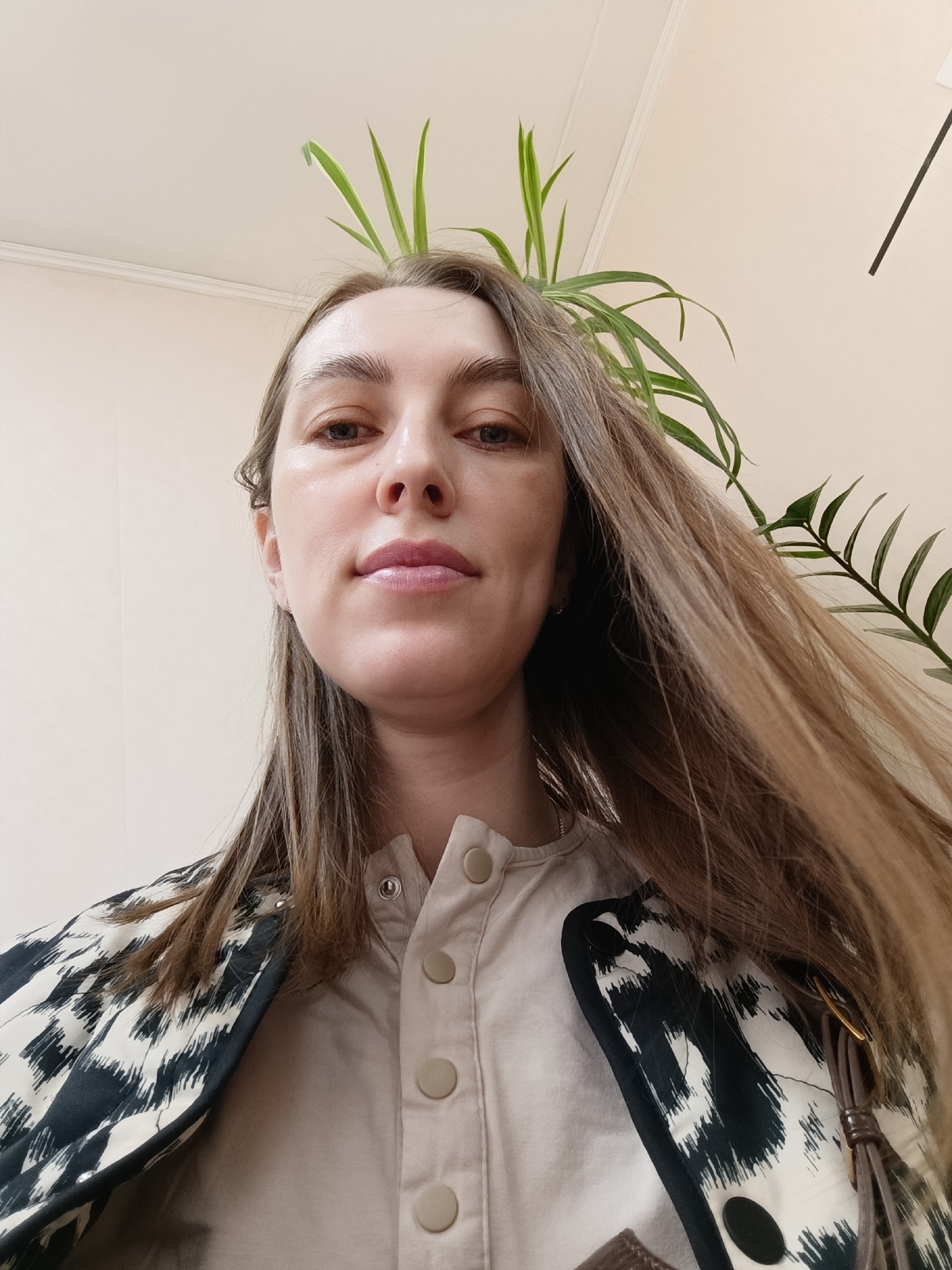 A low-angle, medium shot captures a fair-skinned woman with light brown hair and light-colored eyes. She is wearing a light beige shirt with round buttons and a black and white patterned jacket. Part of a potted green plant is visible in the background, appearing to sprout from her head. The background is a light beige wall with a white ceiling. There is no indication of the city of Chaikovsky, Russia in this image.