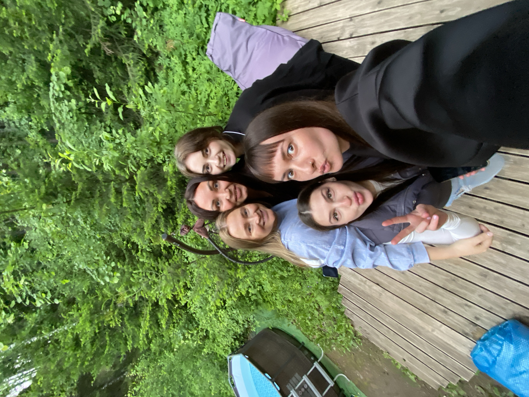 Five young adult females are depicted in a selfie on a wooden deck in Bakhtiyary, Russia. The woman in the immediate foreground, center-right, has dark hair with bangs and performs a puckered 'kiss' facial expression, wearing a black long-sleeved top. To her left, another woman with dark hair gives a peace sign gesture, dressed in a dark vest over a white long-sleeved top and light blue jeans, with her face partially cropped. Behind her, a blonde-haired woman smiles, clad in a light blue long-sleeved shirt. Another woman with dark hair and a visible white collar smiles from behind the blonde individual, wearing a black top. A fifth woman with dark hair is partially visible on the far right, also smiling.
The group stands on a light-colored wooden deck composed of parallel planks, extending across the lower portion of the frame. To the left and slightly below the deck, an above-ground swimming pool with a dark-toned exterior and a light blue patterned interior is visible, featuring an attached ladder. A large, light purple elongated pillow or beanbag rests on the deck behind the women on the right side. A crumpled blue object, possibly a bag, is situated on the deck in the bottom-left corner. The background is dominated by dense, vibrant green foliage, indicating a heavily vegetated, outdoor environment. The scene is illuminated by natural daylight.