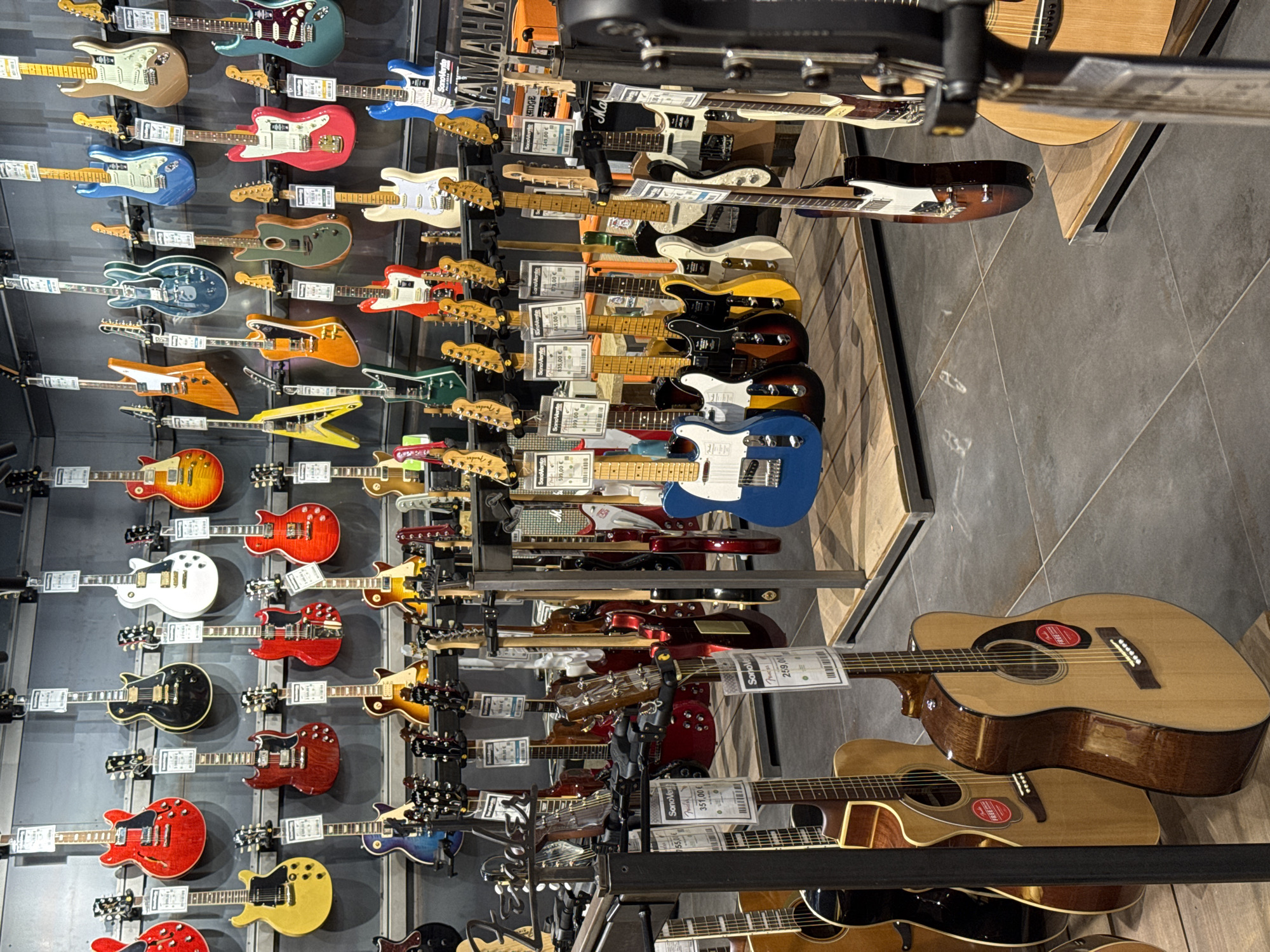 The image shows a retail display of numerous electric and acoustic guitars, predominantly arranged on a metal gridwall display and a wooden shelf. The guitars are displayed at the SonoVente.com showroom location in Palaiseau, France, likely for sale. The guitars are arranged by type and color, with electric guitars mostly on the wall and acoustic guitars primarily on a wooden shelf. Price tags and labels are visible on some guitars. The environment appears to be a well-lit retail space with a tiled floor. The focus is on the presentation of the musical instruments and the orderly arrangement within the store setting.