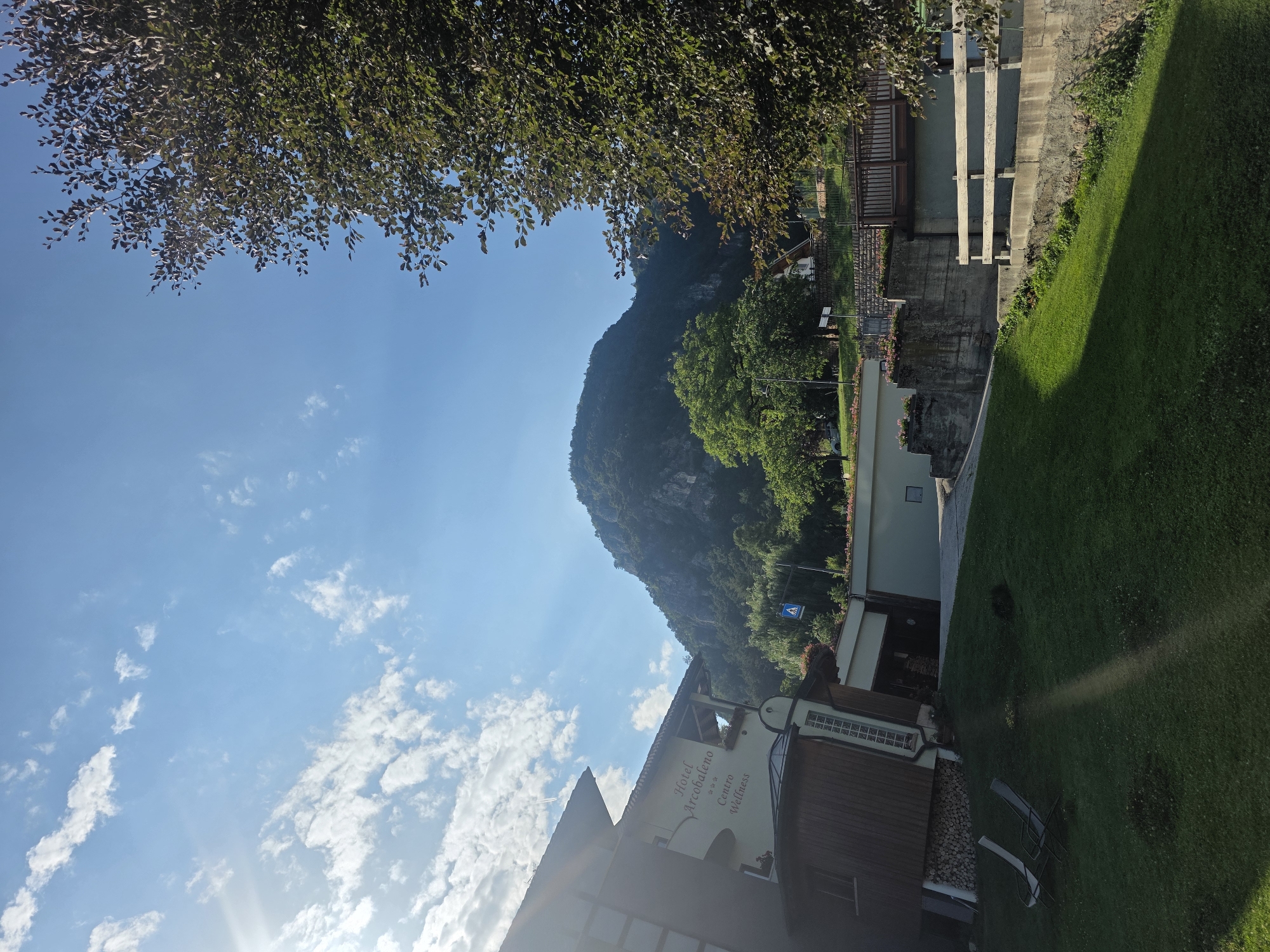 The image depicts a view of Hotel Arcobaleno Centro Wellness in Fai della Paganella, Italy, under a clear blue sky with scattered white clouds. A prominent mountain range with lush green trees covers the background, with the peak shrouded in shadow. In the foreground, a hotel building is visible on the left, featuring a wooden facade and a sign that reads "Hotel Arcobaleno Centro Wellness". Two empty lounge chairs are placed on a well-maintained green lawn in front of the hotel. A paved walkway leads away from the hotel, passing by a concrete retaining wall and a wooden fence. A pedestrian crossing sign is visible on the right side of the image, near the road. The overall scene suggests a tranquil and scenic setting, possibly during daytime on a pleasant day.