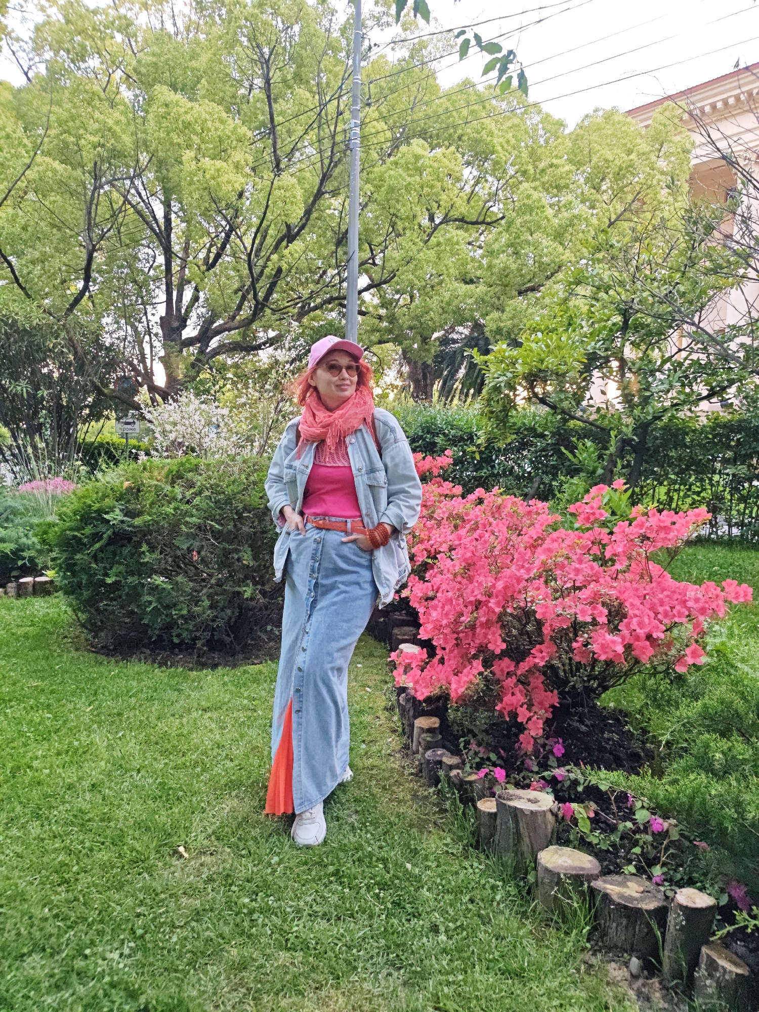 The image shows a woman standing in a park or garden setting. She is wearing a pink hat, sunglasses, a pink top, a denim jacket, a long denim skirt with an orange accent, and white sneakers. She has her hands in her pockets. To her right is a bush with vibrant pink flowers. The background includes greenery, trees, and a building in the distance. The location is Sochi, Russia. The garden includes a trimmed grass area and flowerbed border made from tree trunks.