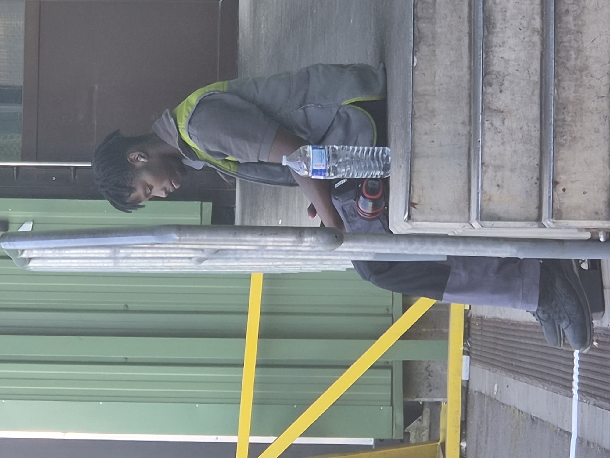 A Black male is sitting on concrete steps outside a building, with his head bowed and eyes closed. He is wearing a grey short-sleeved shirt with a high collar, a grey vest with a lime green collar and trim, and grey pants. He has black dreadlocks and is wearing white wireless earbuds. A partially consumed bottle of water stands upright to his right, next to a dark-colored bag or backpack. To his left, resting on the step below, is a black and red cylindrical object, possibly a speaker or a tool. The background features a tall green corrugated metal wall with a yellow diagonal support beam. A silver metal railing is positioned vertically in front of the man. The environment suggests an industrial or warehouse setting. The scene appears to be captured in Leudelange, Luxembourg, given the provided context of the image's origin.