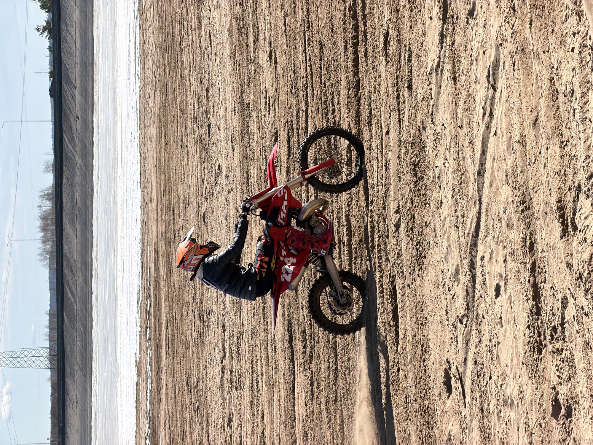 A motocross rider is performing a wheelie on a dirt track in Chaikovsky, Russia. The rider, wearing an orange helmet, black jacket, and multi-colored pants, is on a red dirt bike with the number 224 on its side. The track is sandy with visible tire tracks, and a snow-covered area with a concrete barrier and distant power lines are in the background. The sky is light blue.