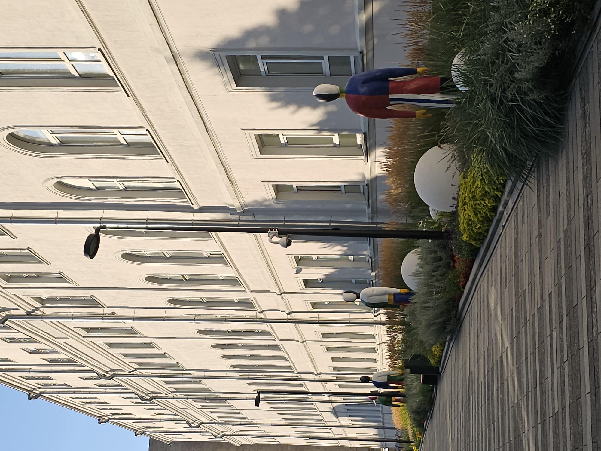 The image captures a sunlit pathway leading towards a light-colored building with numerous windows. Several colorful, abstract statues of people are positioned in a landscaped area alongside the pathway. The pathway itself is paved with rectangular stones. In the background, the building has a classical architectural style with arched windows on its upper floors. The statues are abstract in design and appear to be made of a solid material, with distinct color blocks. The landscaping includes grasses and other low-lying plants. A street lamp is visible on the pathway. The scene appears to be an outdoor public space in a city. This scene is located in Moscow, Russia, specifically at the National Research University Higher School of Economics (HSE) at 20 Myasnitskaya Street.