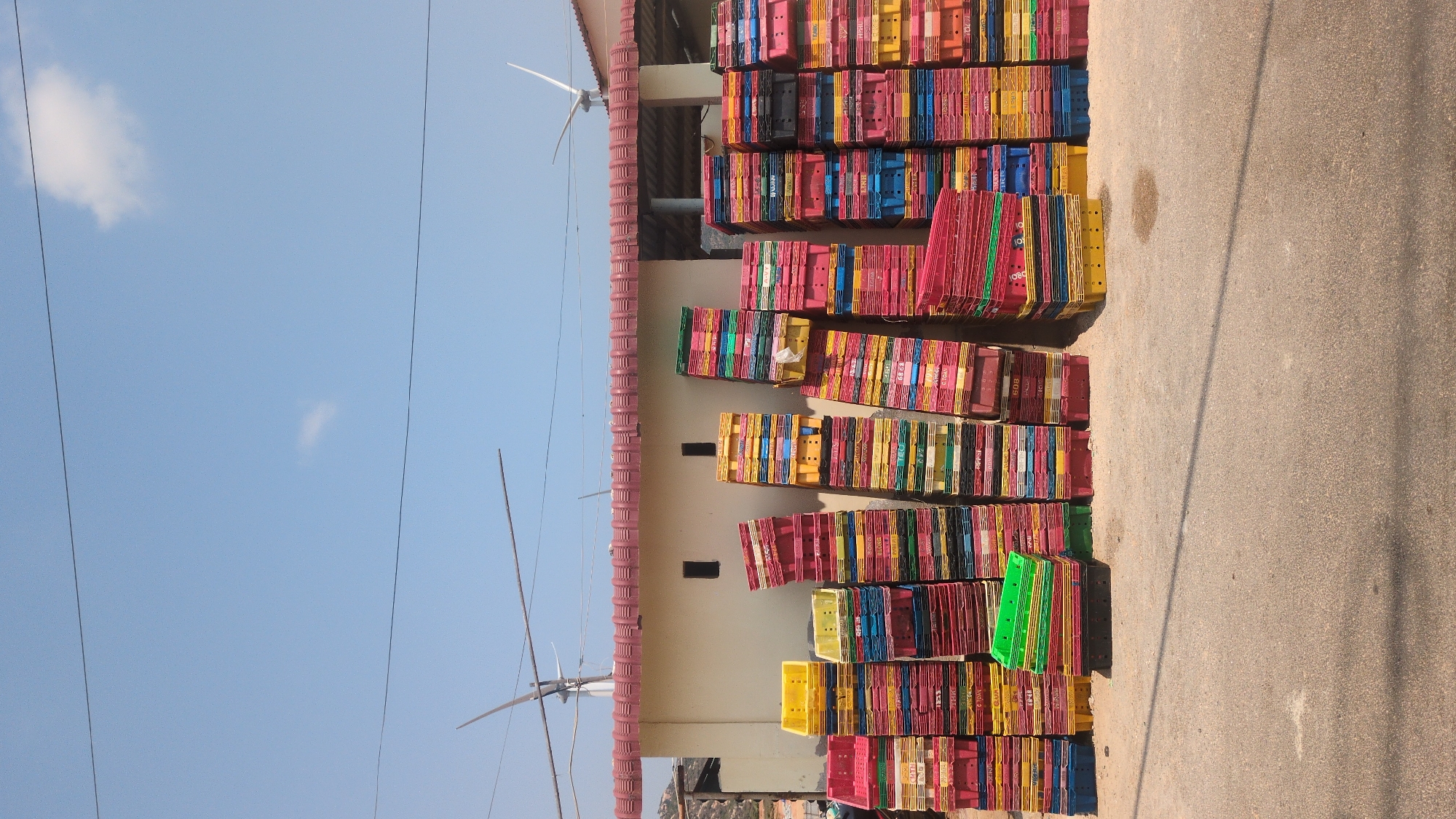 A large pile of stacked colorful plastic crates sits beside a building in Phước Dinh, Vietnam. The crates are predominantly red, yellow, green, and blue, and stacked in tall columns. The building has a pale-colored exterior and a red roof. A paved area stretches to the right of the crates, appearing to be a roadway or parking area. Electrical wires span across the top of the frame against a clear blue sky. A small wind turbine is attached to the building's roof.