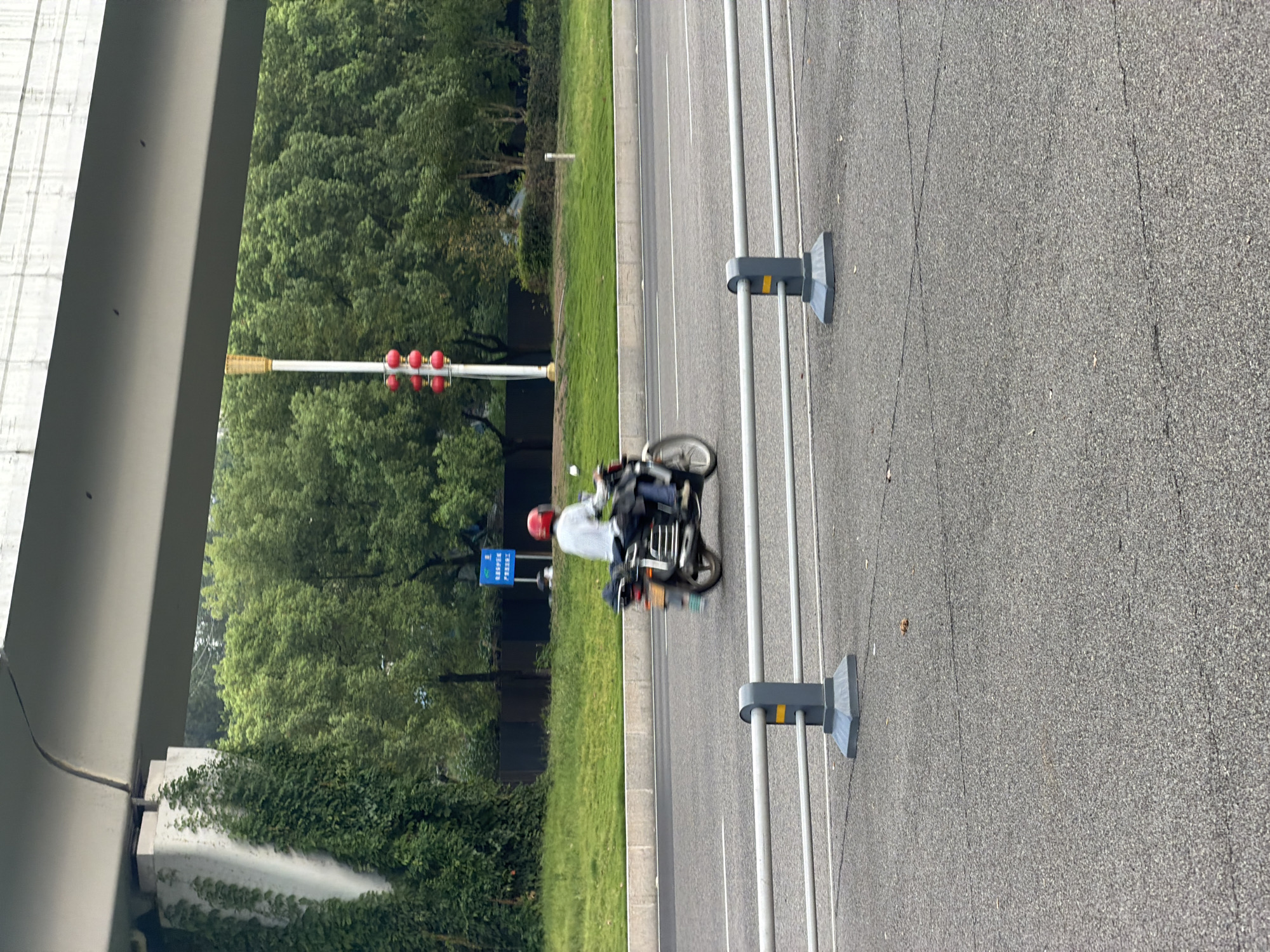 A person wearing a red helmet and a light-colored shirt rides a motorcycle on a paved road in Jiangjin, China. The motorcycle is black and has a silver rack on the back. The person is in motion, as indicated by the blur in the image. In the background, there are trees, a traffic light with red bulbs, and a sign. A concrete overpass runs across the top of the image. The road is divided into lanes by white lines, and a metal guardrail runs along the side.