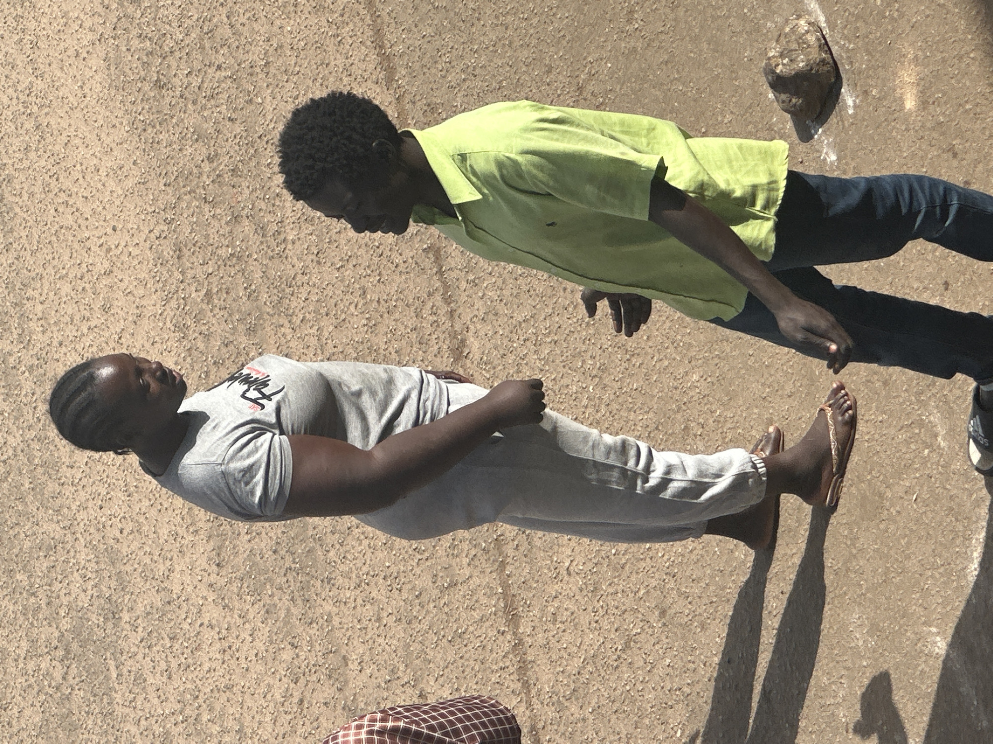 The image shows two individuals standing on a dusty, unpaved surface, likely a street in Bukuru, Nigeria. To the left, a woman with dark skin and braided hair stands facing right, wearing a gray t-shirt with a graphic print and light gray sweatpants. She is wearing flip-flops. To the right, a man with dark skin and short, curly hair faces the woman, wearing a short-sleeved, lime green collared shirt and dark blue jeans. His hands are open and appear to be gesturing. The environment is a rough, textured ground with some scattered pebbles and a rock to the right of the man. The lighting suggests it is daytime, with noticeable shadows cast by the individuals. There is also a small portion of another person visible on the far left, wearing a brown and white checkered garment.