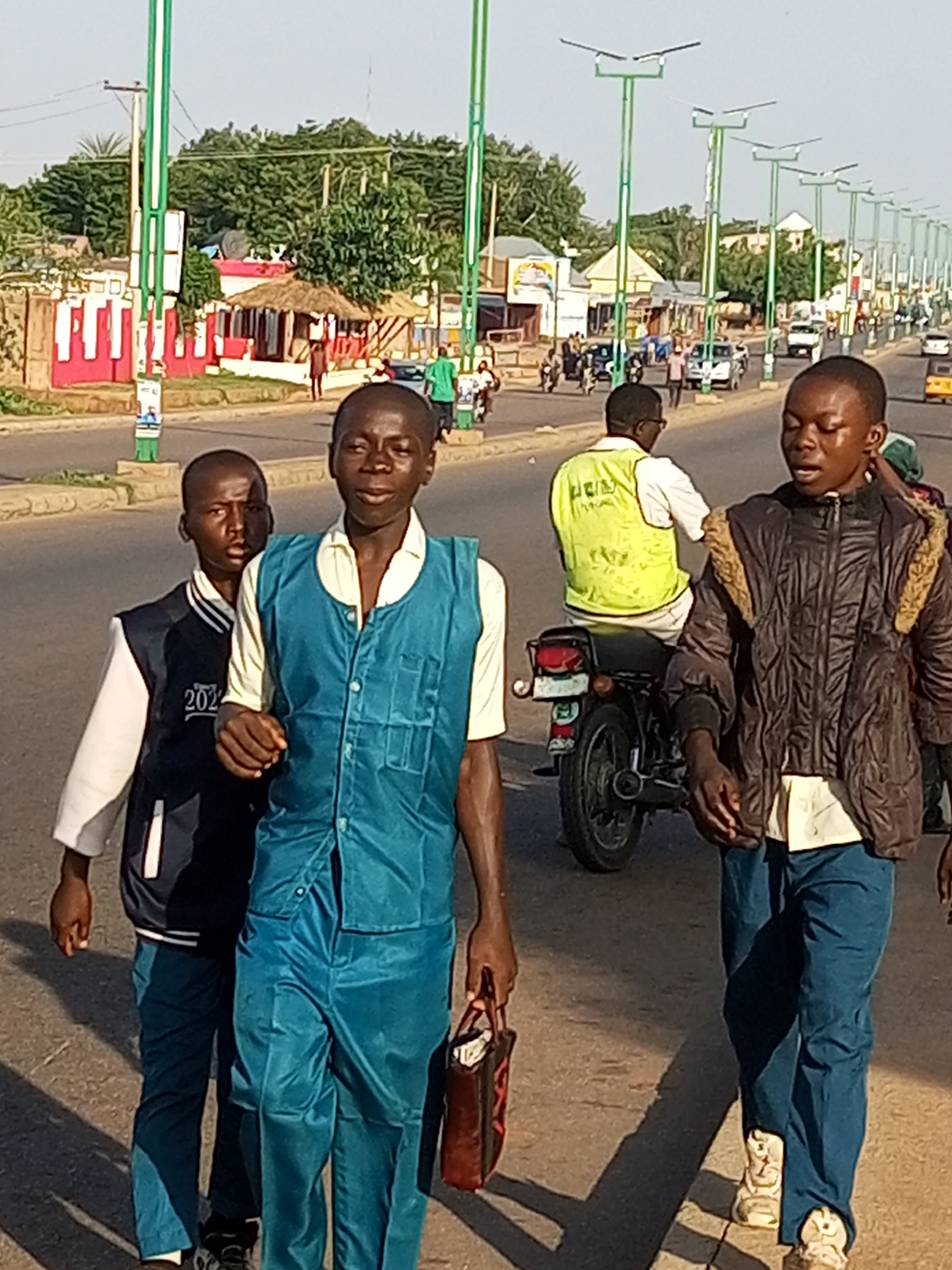 Three young Black males are walking on a paved road in Garko, Nigeria. The male in the foreground on the right wears a brown jacket with fur trim and a light-colored shirt and blue pants. He carries a red bag in his right hand. The male in the middle foreground wears a teal vest over a light-colored shirt and teal pants. The male on the left foreground wears a dark jacket with white sleeves and a "2024" inscription and dark pants. In the background, a man in a bright yellow vest is seated on a motorcycle. Further in the background, there are buildings with red and white facades, thatched roofs, trees, and streetlights. Traffic, including cars and motorcycles, is visible on the road. The sky is clear and bright.
