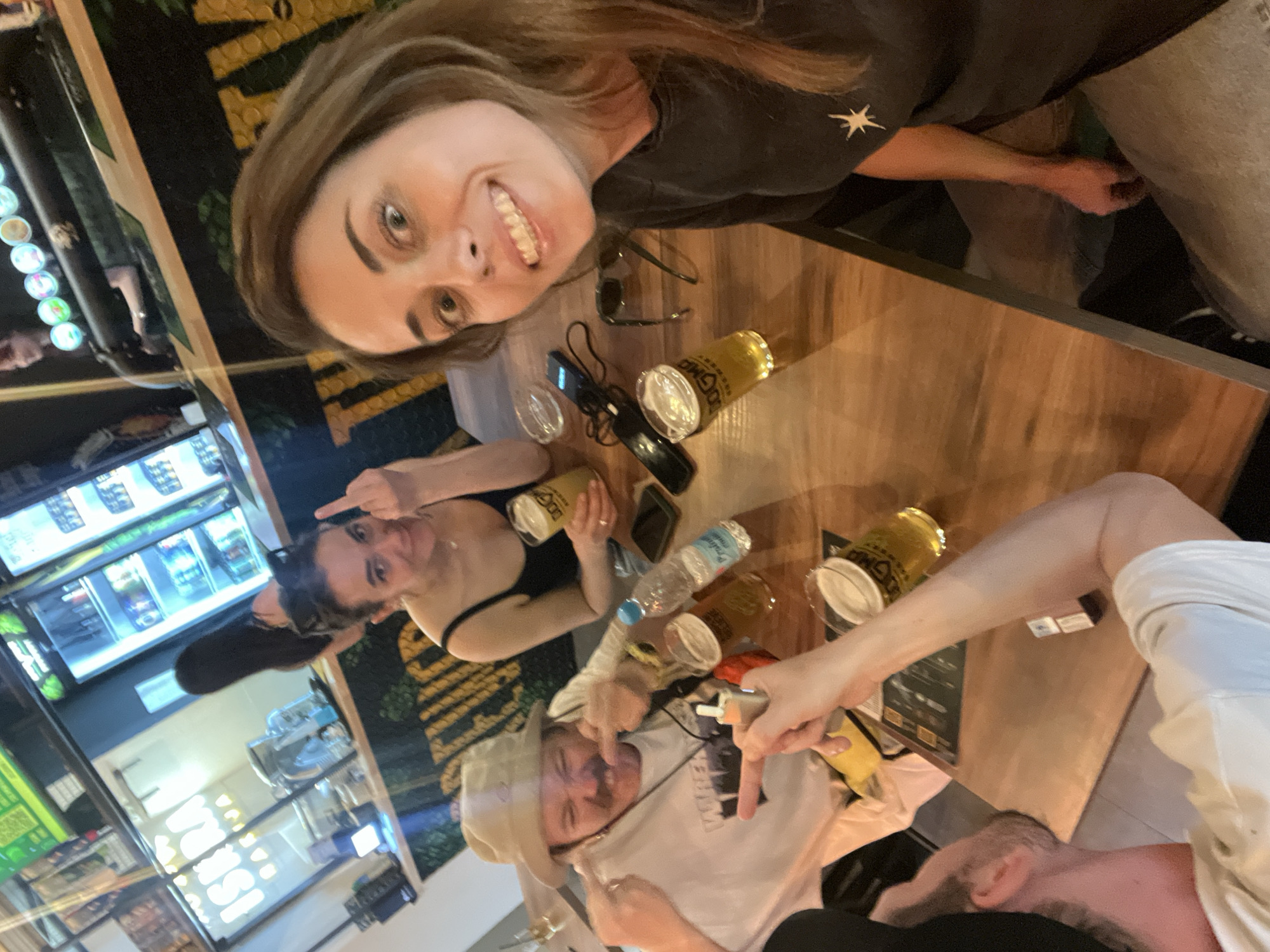 The image captures a group of four people sitting around a wooden table at what appears to be an outdoor bar or restaurant. The photograph is taken from a tilted angle, with a woman at the top of the frame smiling directly into the camera. There are numerous bottles of beer and a water bottle on the table. One woman is seen gesturing with her middle finger raised. Another person is pointing at the individual wearing a white cowboy hat, who is also gesturing with their middle finger. The background reveals a refrigerated display case filled with beverages, suggesting a commercial establishment. The location is Belgrade, Serbia.