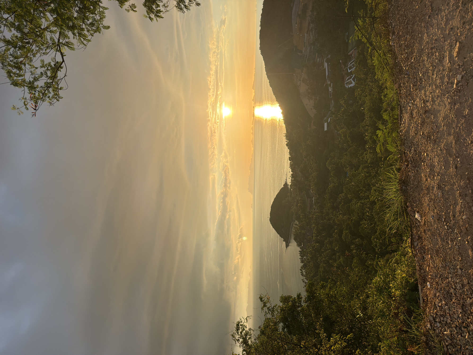 A high-angle, full outdoor shot captures a sunset over the ocean, viewed from a hillside overlooking a lush, tropical landscape. The sun, a bright yellow orb, is positioned in the upper-middle of the frame, casting a golden reflection across the water. Islands are visible in the distance, silhouetted against the horizon. The foreground consists of a dirt path with scattered gravel and vegetation. In the midground, dense green trees and foliage cover rolling hills, with some buildings and construction visible among the trees. The sky is a blend of soft oranges, yellows, and grays, with clouds stretching across the upper portion of the image. The overall scene depicts a serene coastal environment during the golden hour.