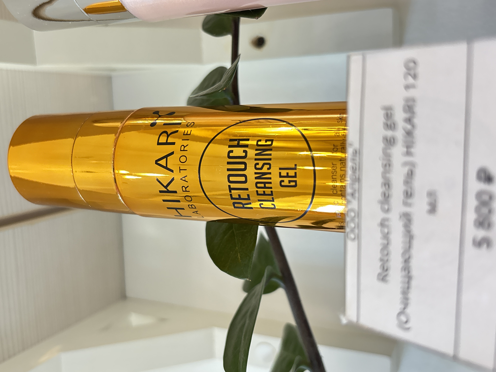 An eye-level, medium shot captures a golden bottle of HIKARI LABORATORIES RETOUCH CLEANSING GEL, positioned on a shelf with a white label in the foreground. The bottle is metallic gold with black lettering. The label displays "Retouch cleansing gel (Очищающий гель) HIKARI 120" and the price "5 800 ₽". To the left of the bottle, a green plant with large leaves is visible. The background is a clean, white wall. The overall lighting is bright and even, suggesting an indoor setting like a store. The image does not provide any information about a specific city or country in Russia.