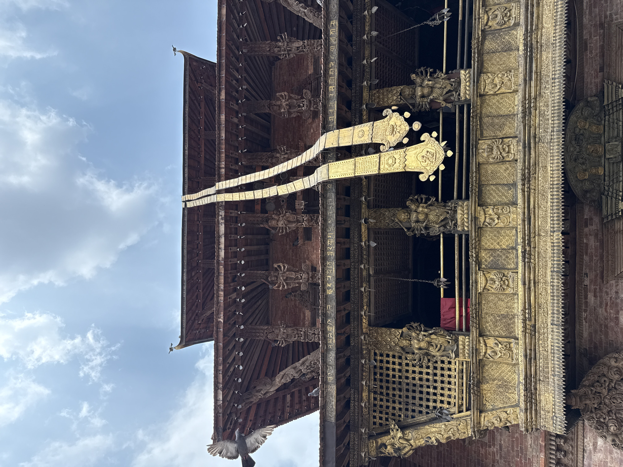 A low-angle shot shows a corner of a multi-tiered building in Lalitpur, Nepal, featuring a brown wooden framework with carved figures, a golden facade, and script etched on vertical beams. A bird is captured mid-flight at the bottom of the frame, against a backdrop of a blue sky with scattered clouds. The ornate, golden facade has a lattice window with a glimpse of red fabric inside.
